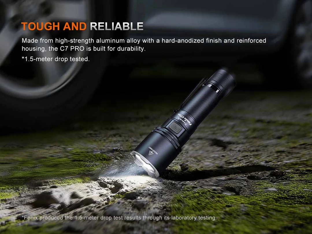 Durable black flashlight on mossy ground after a 1.5‑meter drop test, car tire faintly in the background.
