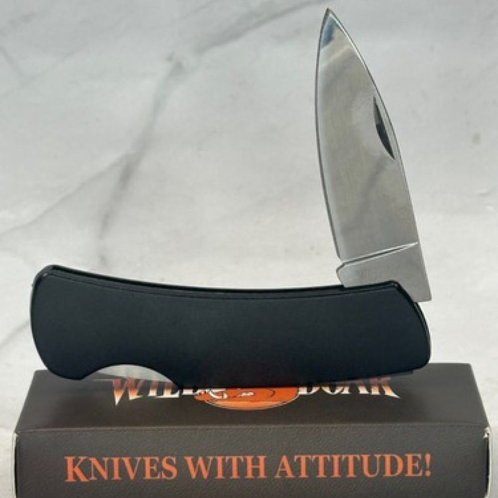 Folding knife with a black ergonomic handle resting on a box that reads 'KNIVES WITH ATTITUDE!'. The blade is partially open, showing a sleek silver finish.