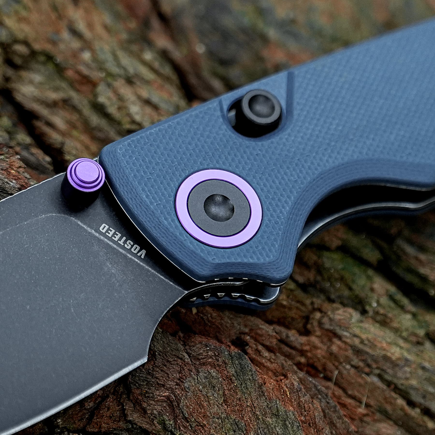 Close-up of a folding knife handle with purple hardware on a textured gray grip, blade partially visible against a wooden background
