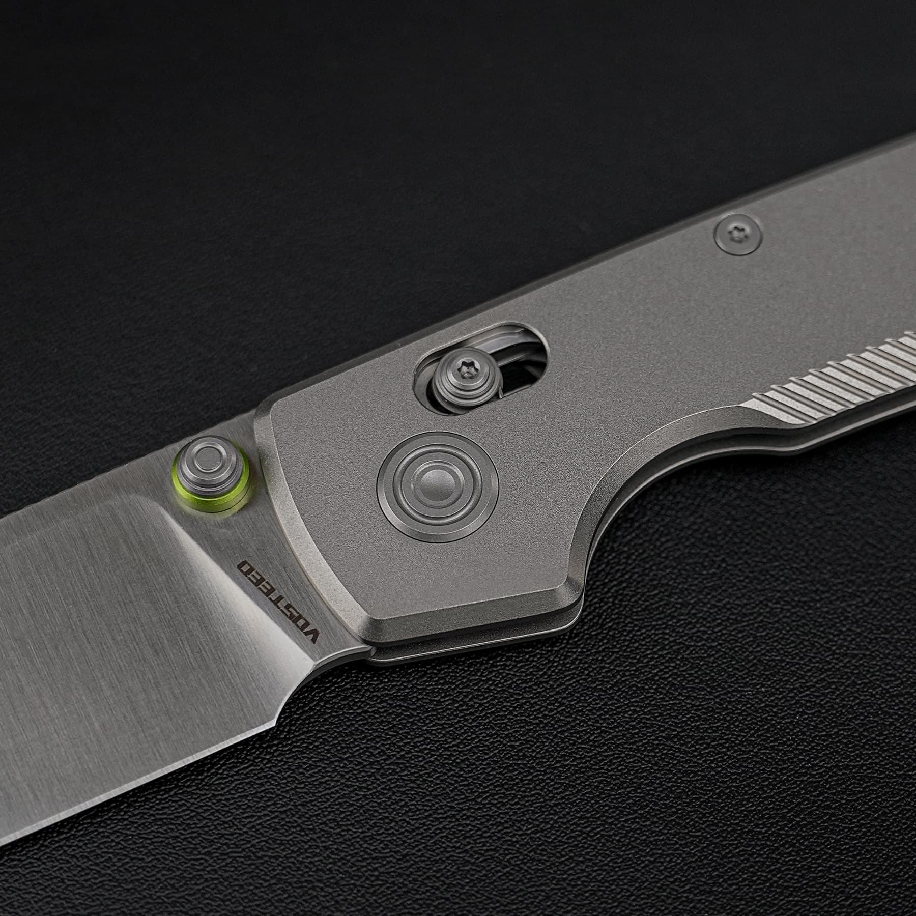 Close-up of a metallic folding knife handle with screws, a pivot, and a green accent on a black textured background.