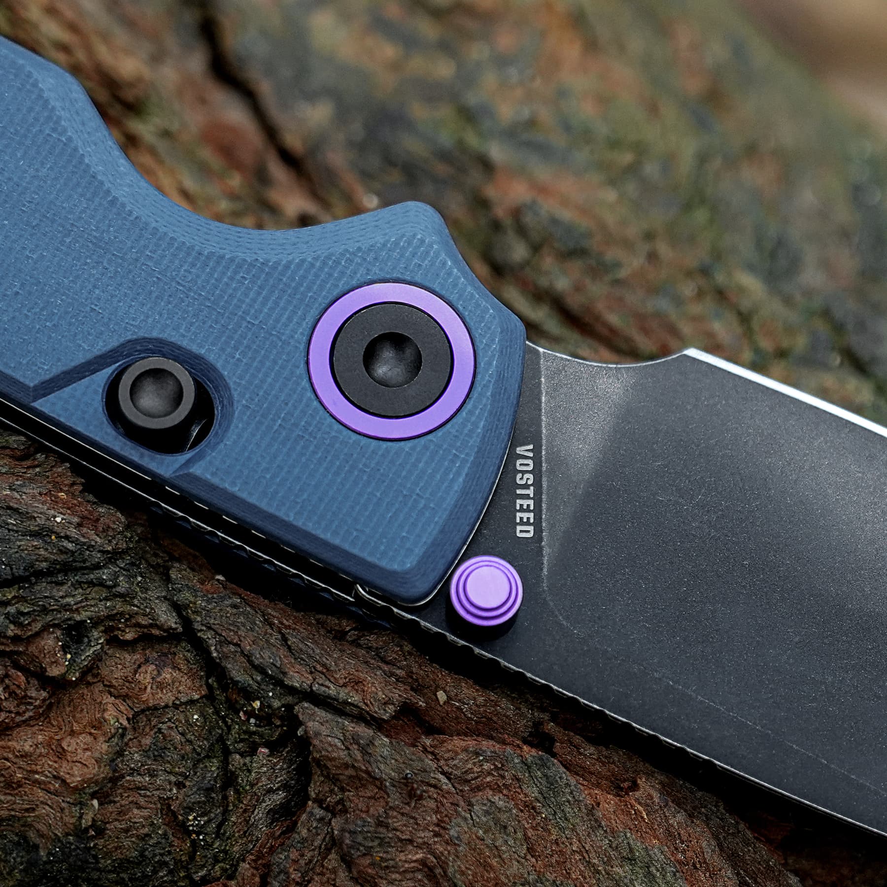 Close-up of a blue folding knife with purple pivot hardware resting on rough bark; blade partially exposed with a black edge and purple accents