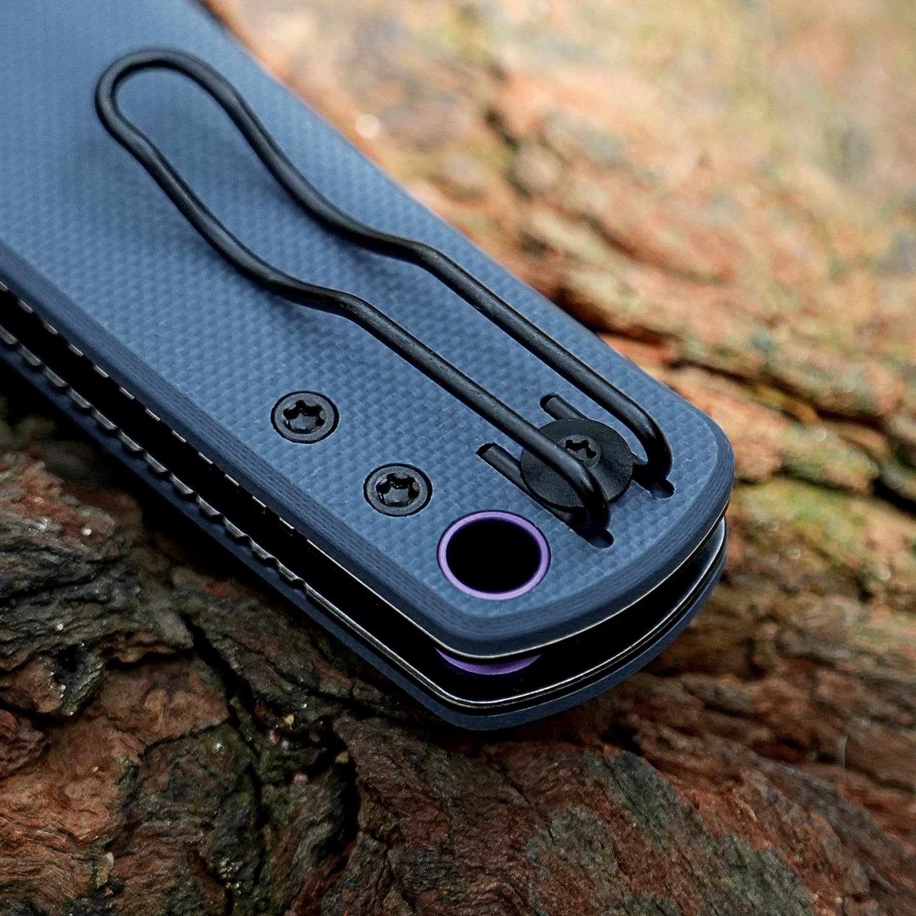 Close-up of a blue textured multitool with a black curved lanyard clip resting on rough tree bark.