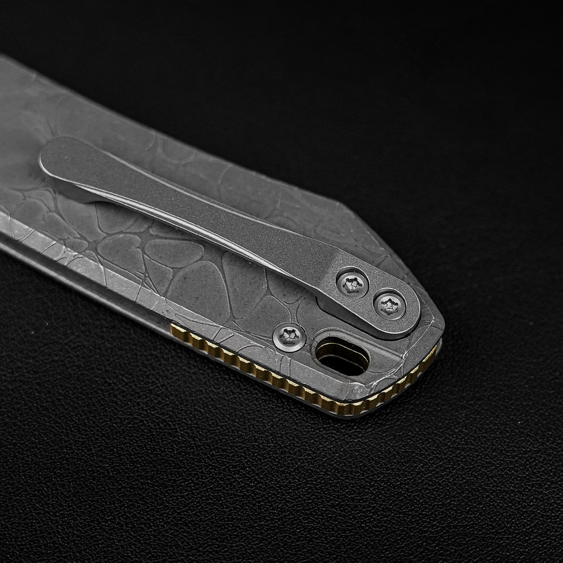 Close-up of a textured gray folding knife with gold-toned serrated edge against a black background as body housing shows screws and design
