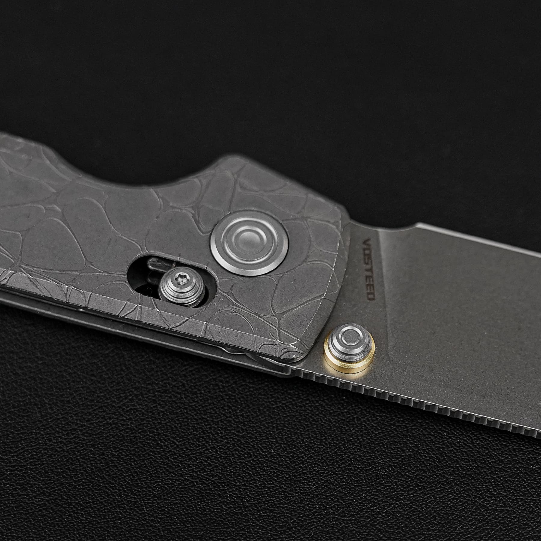 Close-up of a textured gray folding knife handle with a round pivot and exposed screws, resting on a black leather-like surface.