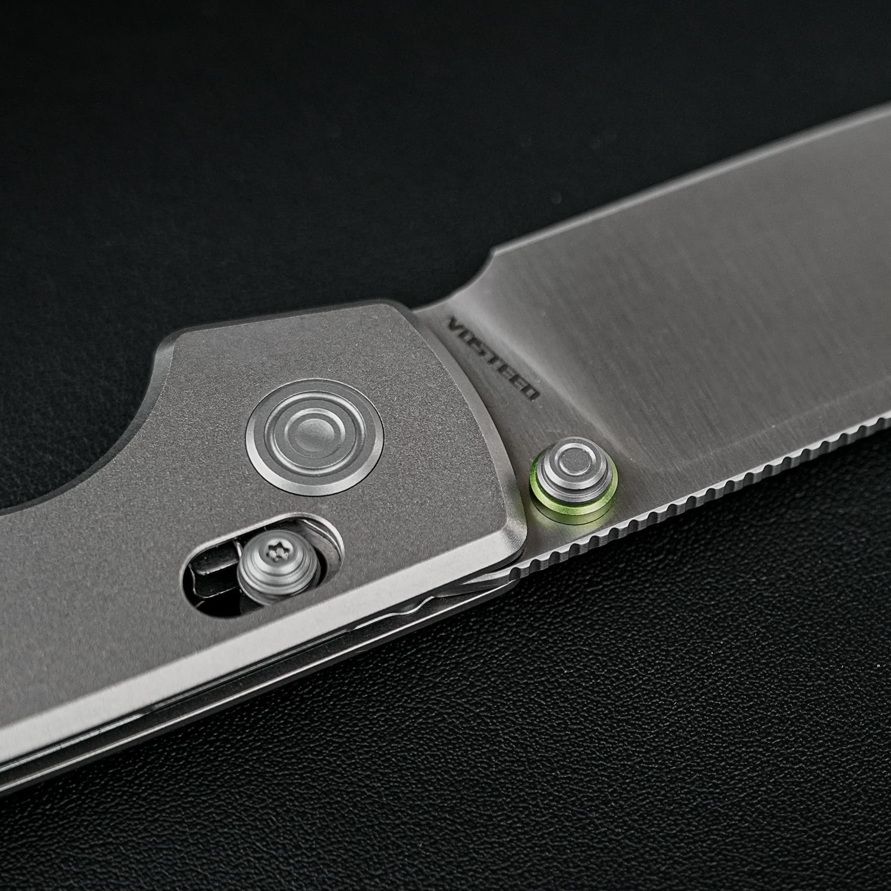 Close-up of a grey folding knife with exposed blade and pivot screws on a black textured background.