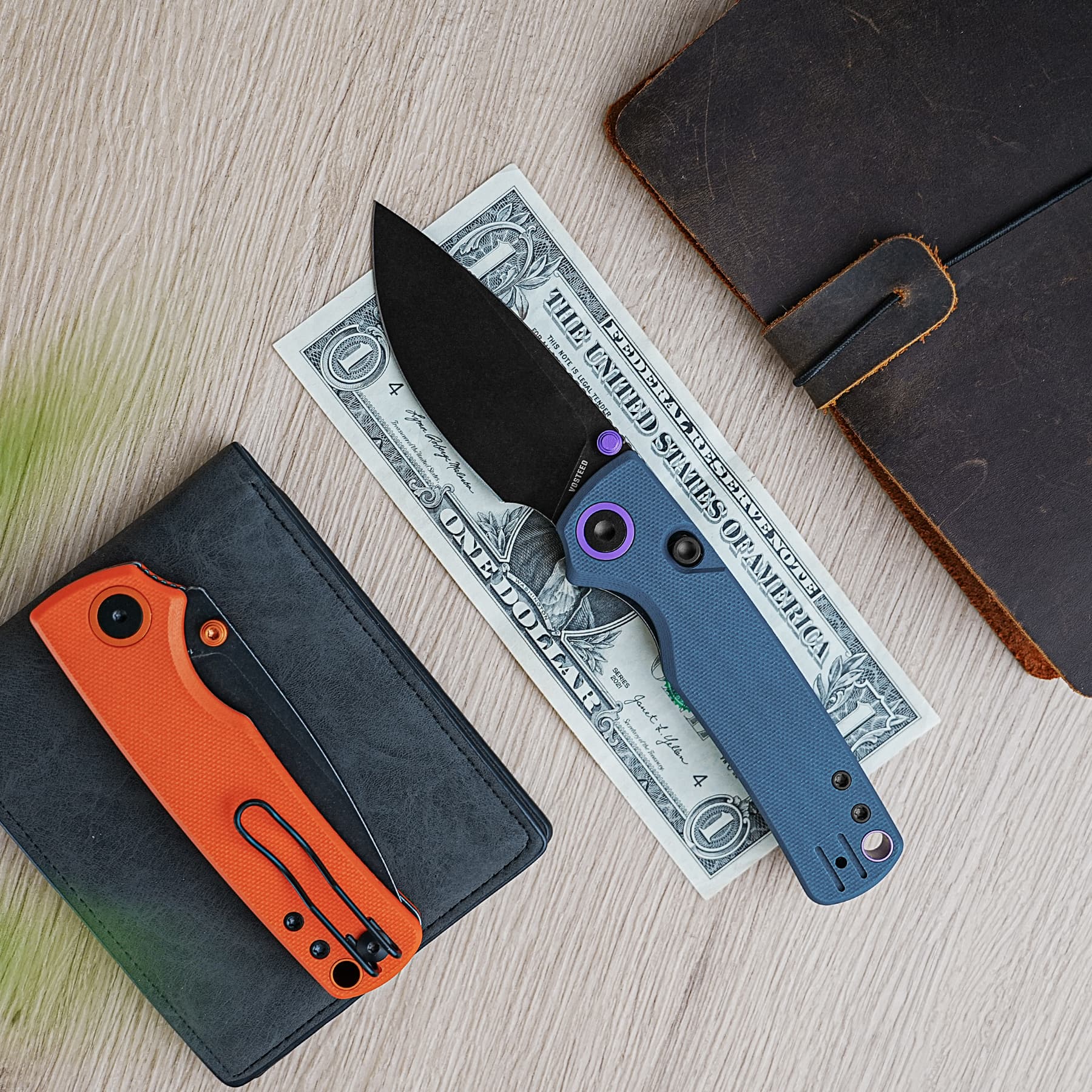 Flat lay of a one-dollar bill tucked under a black knife blade with an orange handle, beside a blue knife and a dark wallet on a wooden surface.