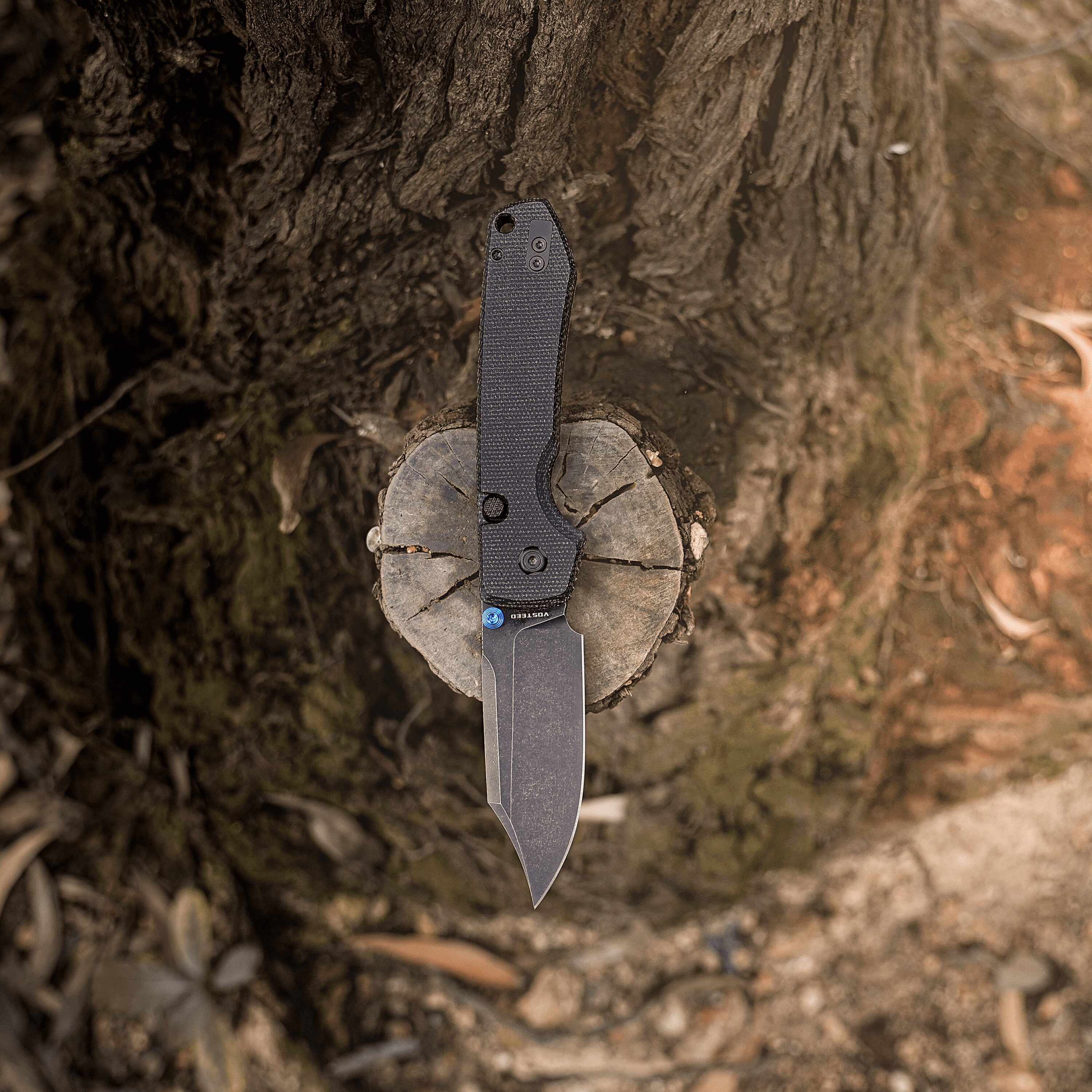 A dark, textured knife embedded in a tree stump, blade vertical and strapped to a worn handle in dirt and roots.