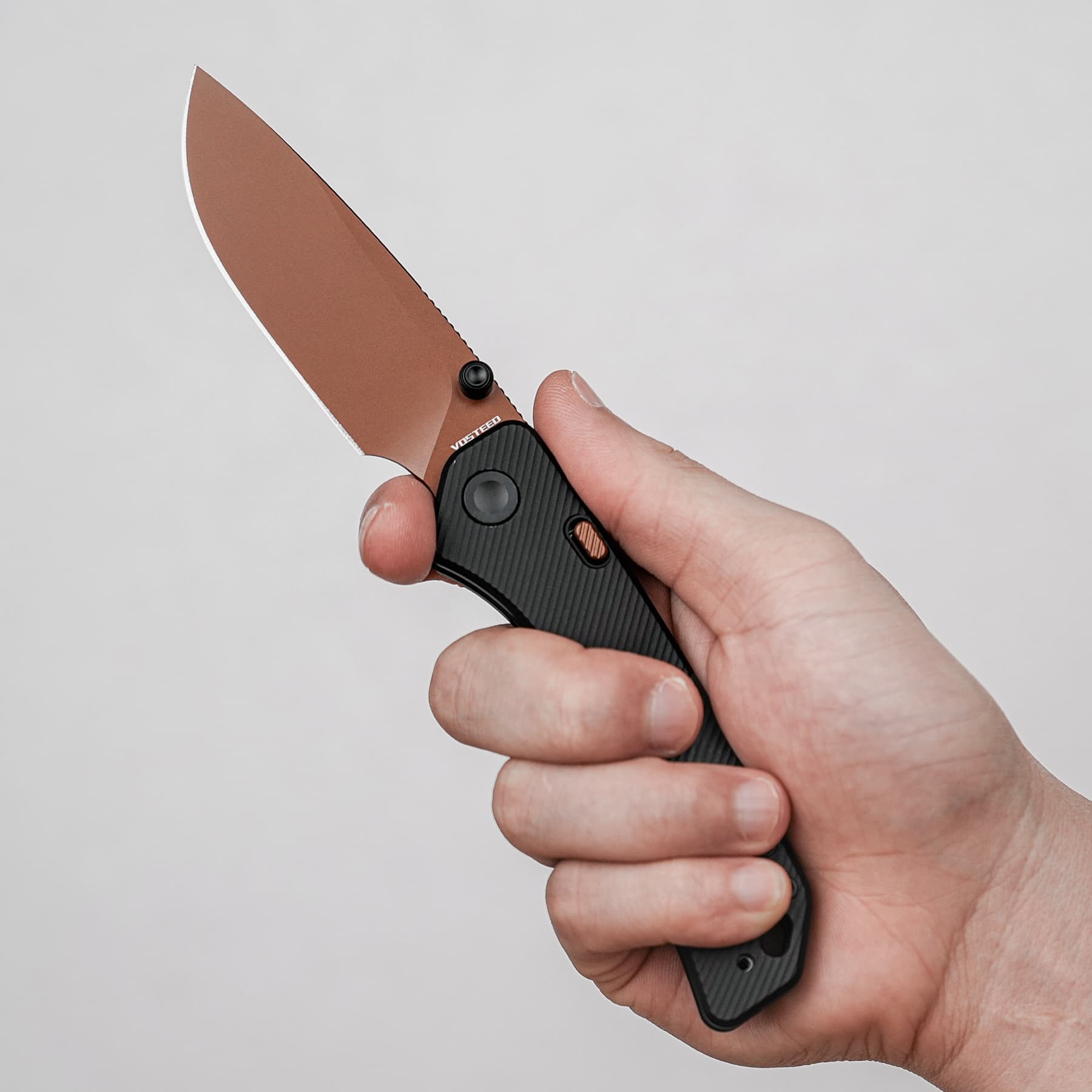 Hand gripping a brown-toned folding knife with a black textured handle and a sharp blade edge visible in the foreground.