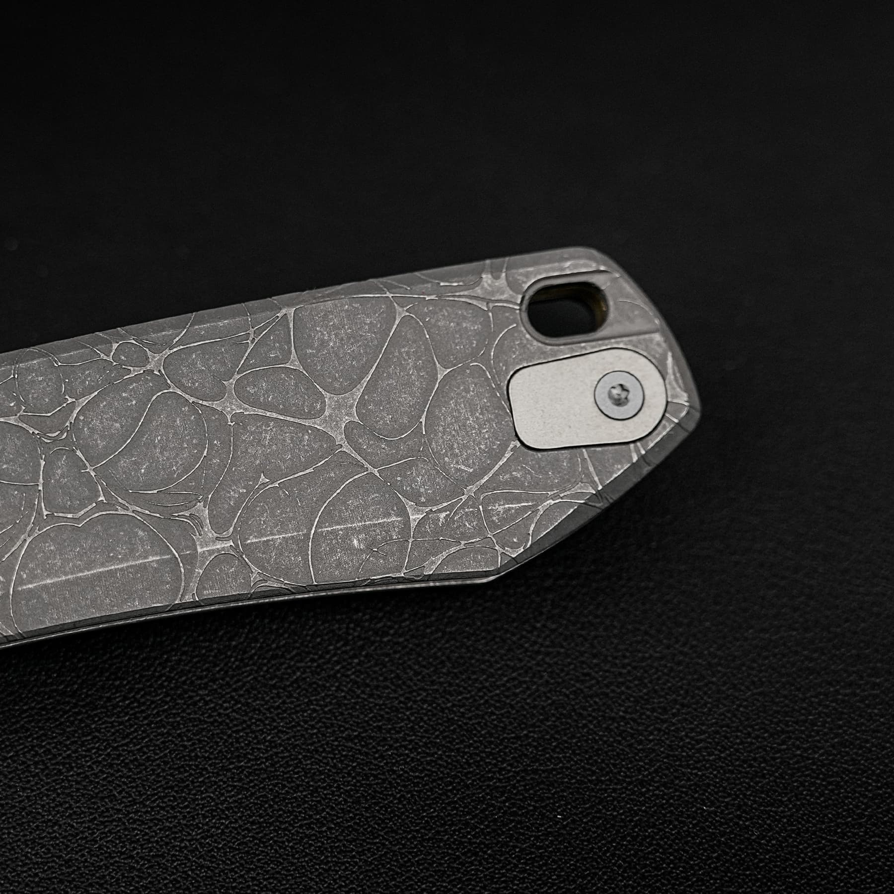 Close-up of a silver folding knife handle with a textured, marbled pattern against a dark background.