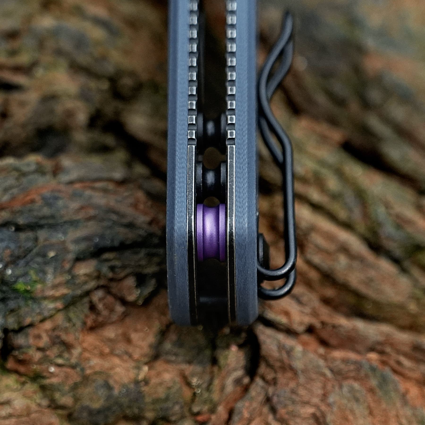 Close-up of a black metal component with two rows of small square teeth facing inward, revealing a purple cylindrical piece in the center against a rocky background.
