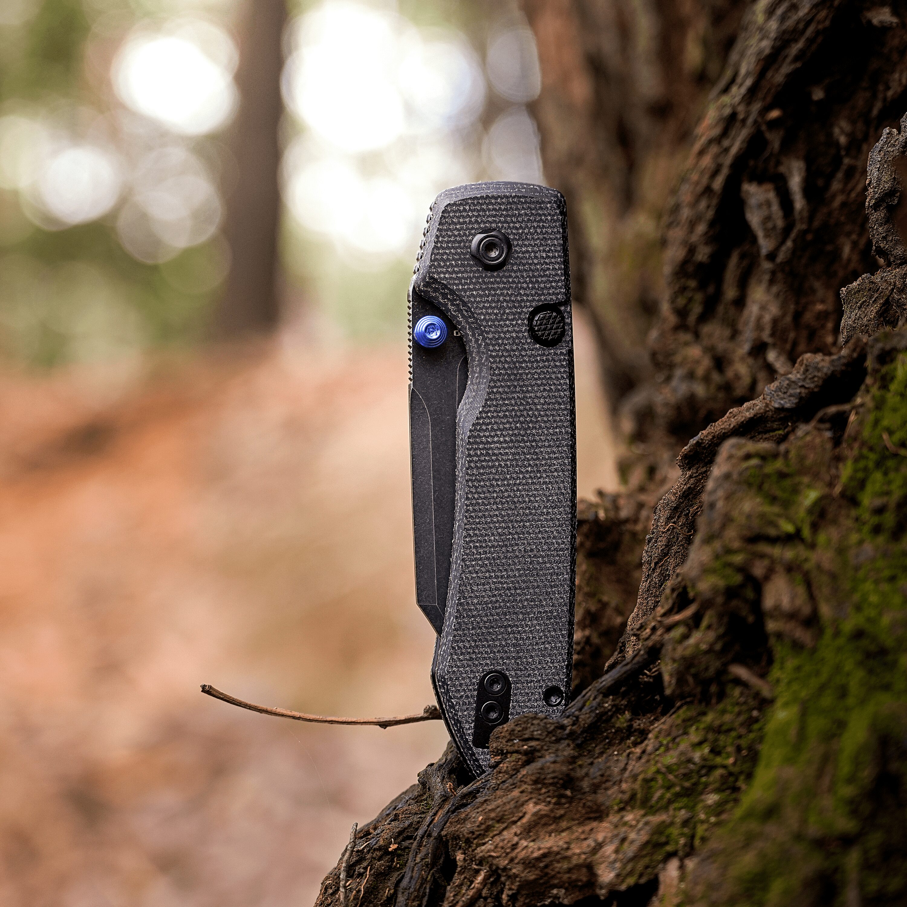 Close-up of a black textured knife blade embedded in a tree trunk in a forest setting.