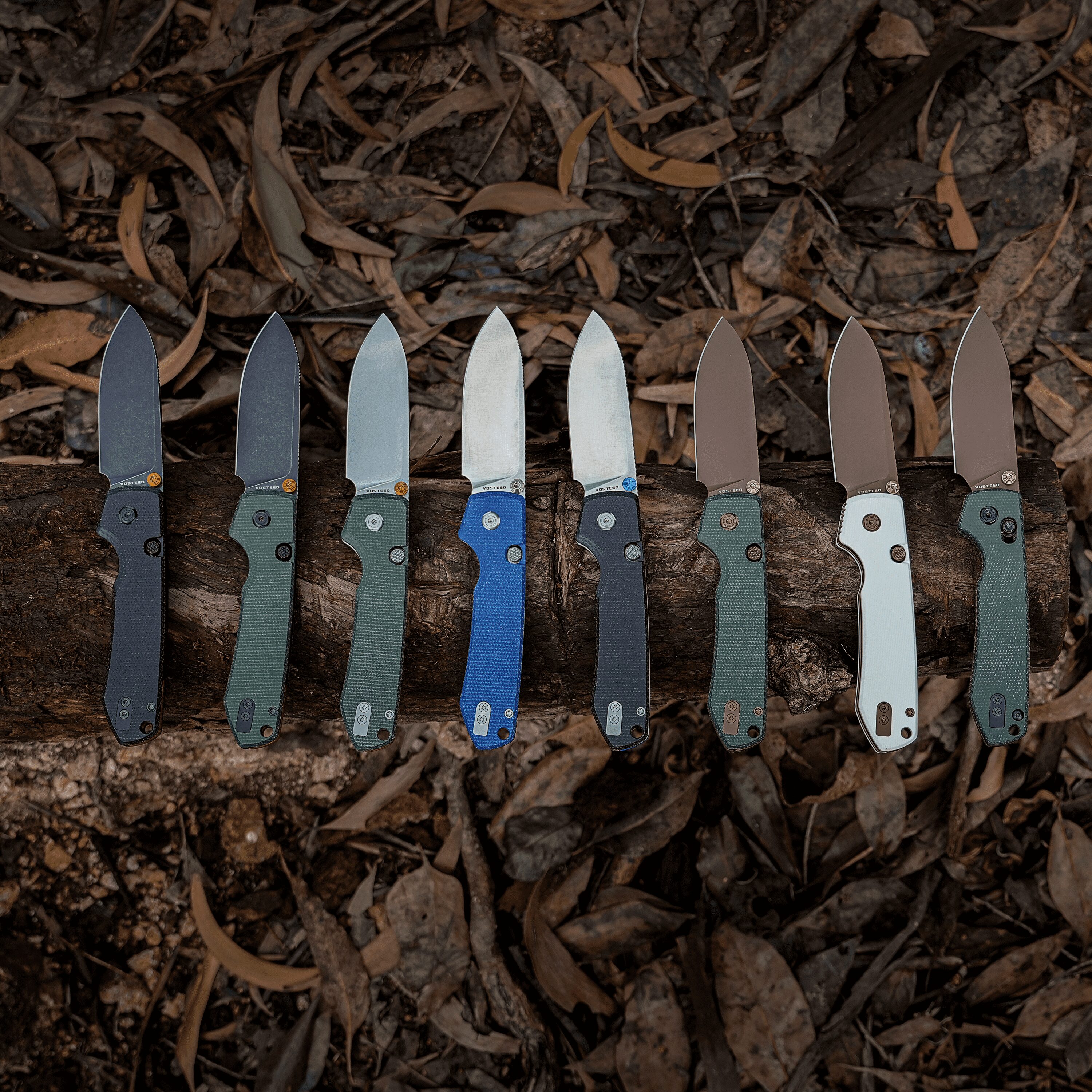 Row of folding knife blades with colored handles laid flat on dirt and fallen leaves.
