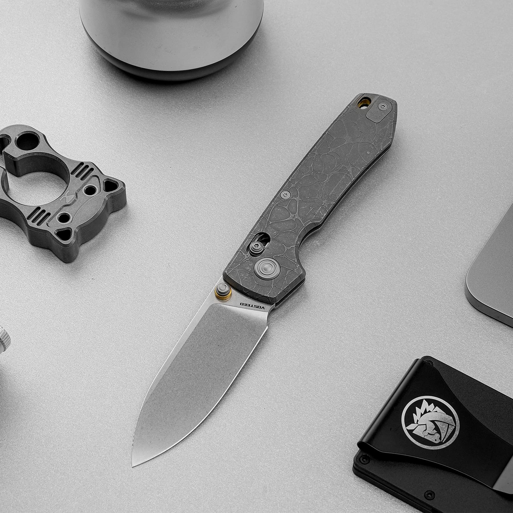 Folding knife with cracked gray handle lying diagonally on a gray work surface, blade extended; a black case and tool component are nearby.