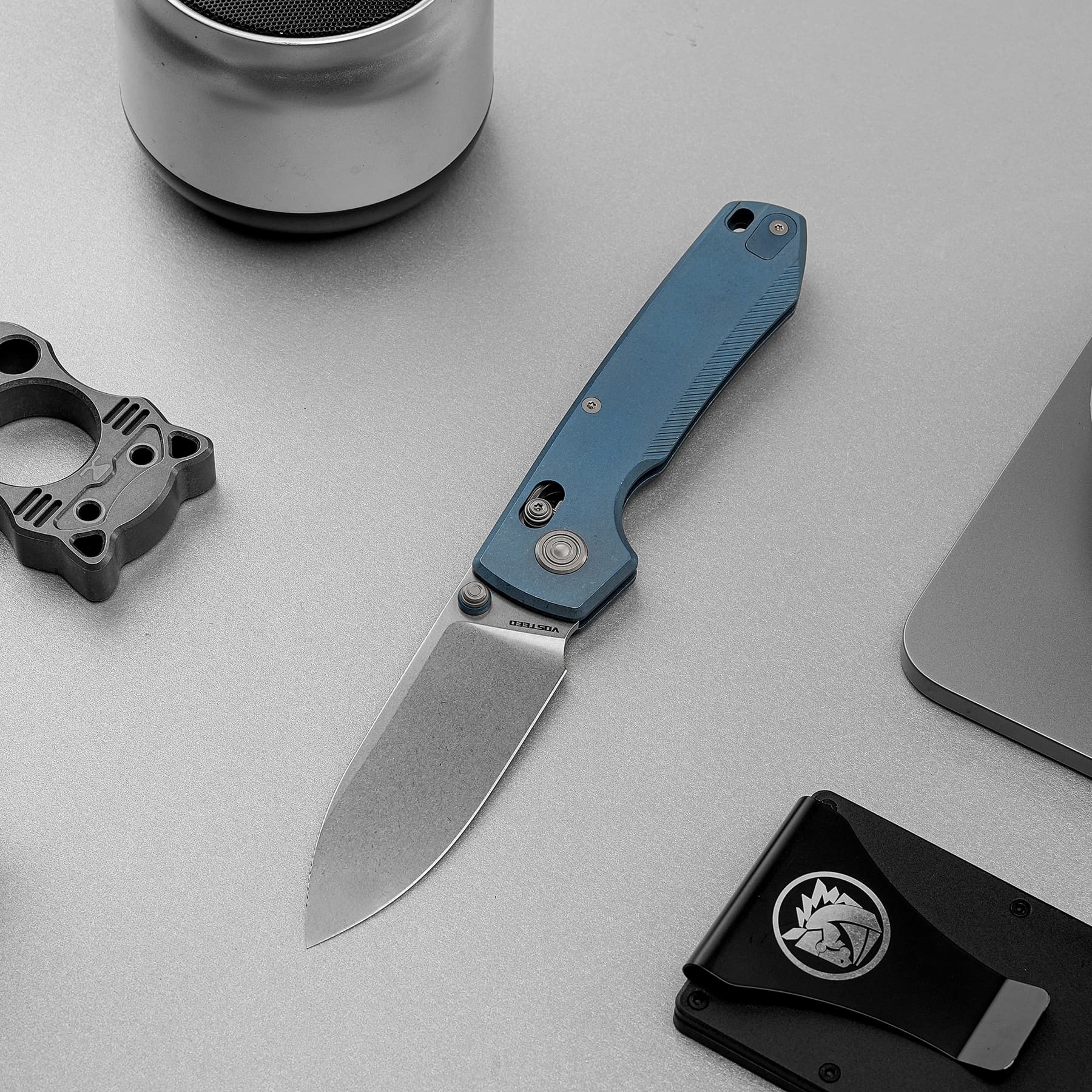 Blue-handled utility knife laid on a gray work surface beside a metal gadget and a black smartphone, with a mechanical part in the lower-left corner.