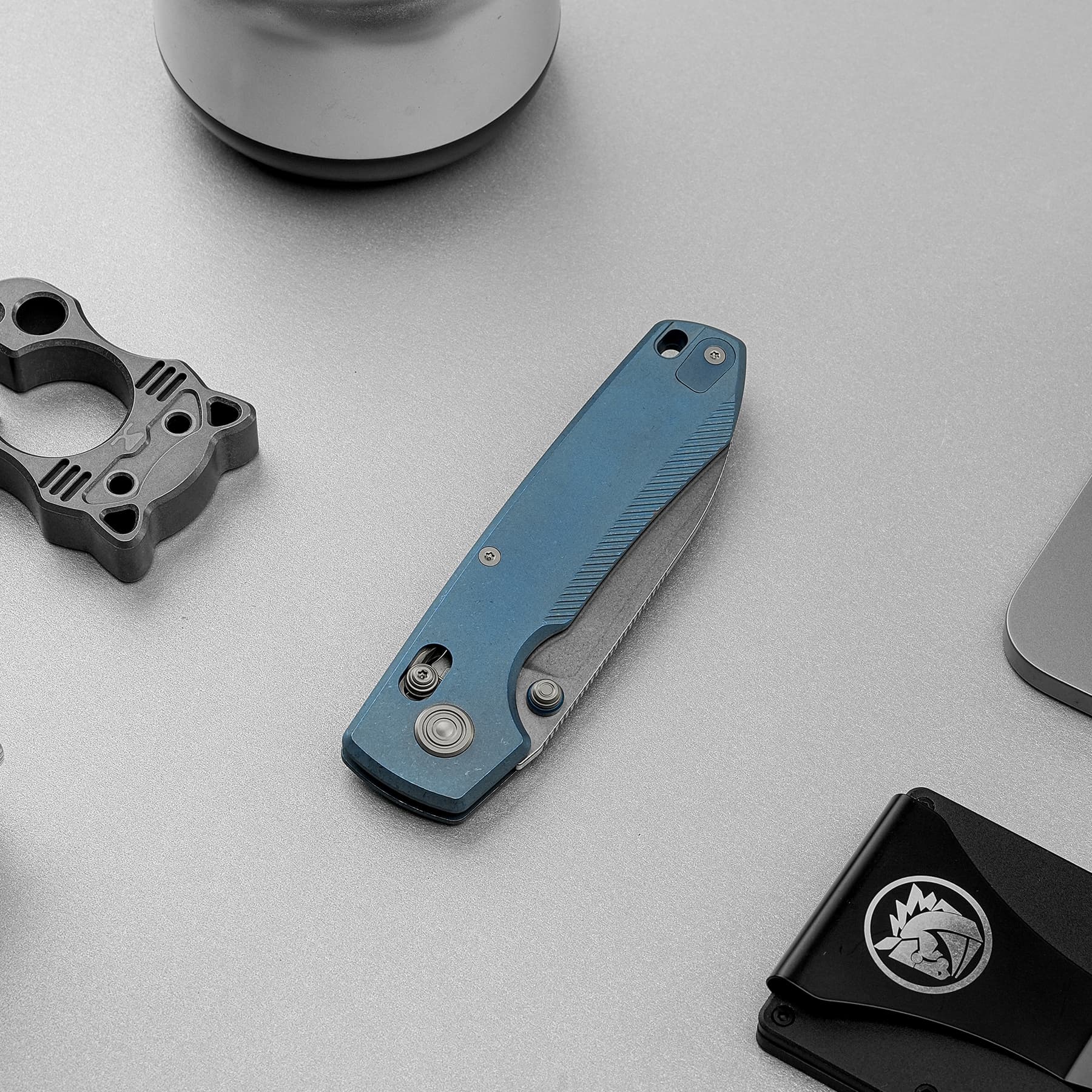 Blue folding knife resting on a gray surface next to a metallic component and a black gadget with a logo nearby.