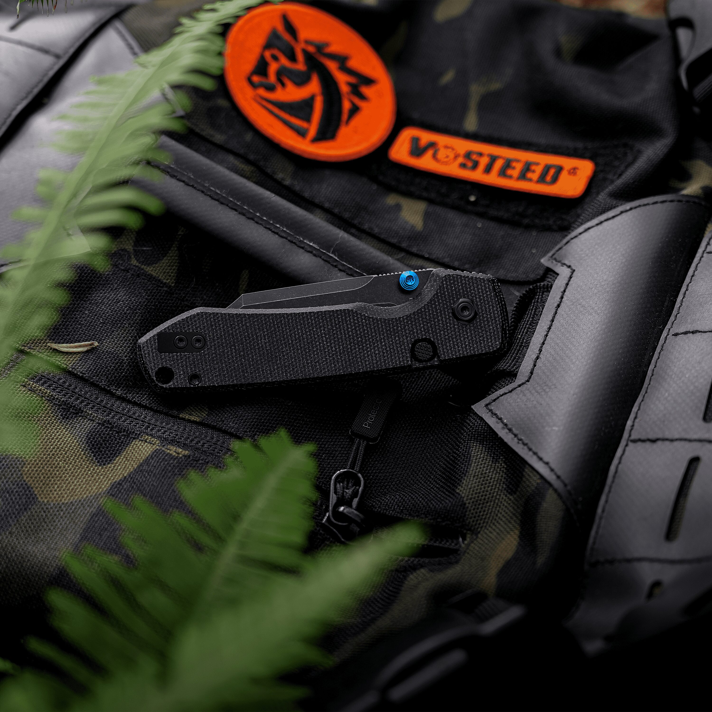 Close-up of black camouflage fabric with an orange circular logo patch and an orange 'VOSTEED' label, partially obscured by green fern leaves in the foreground.