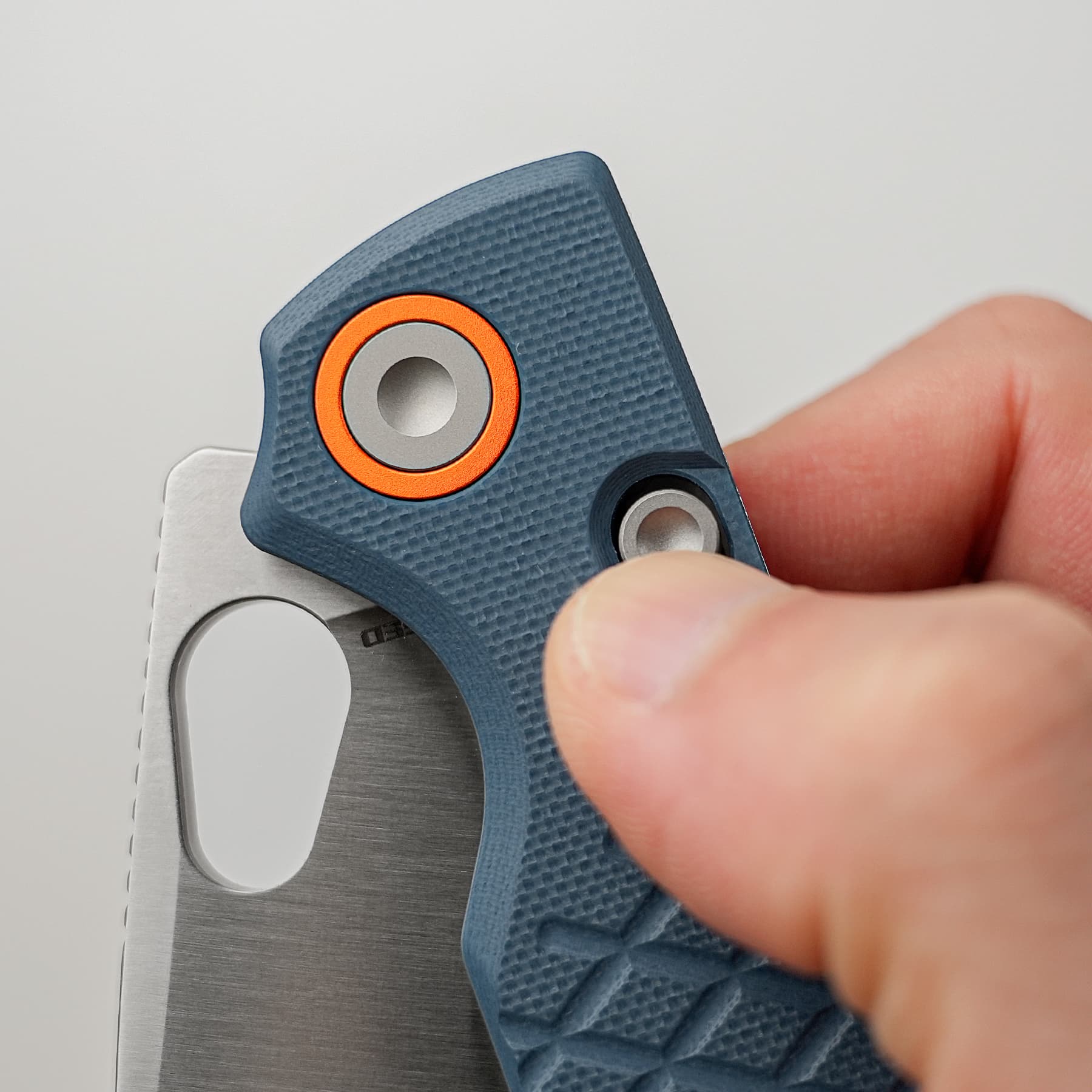 Close-up of a blue textured multitool being held, showing a sharp blade and orange-ringed pivot near a finger grip.