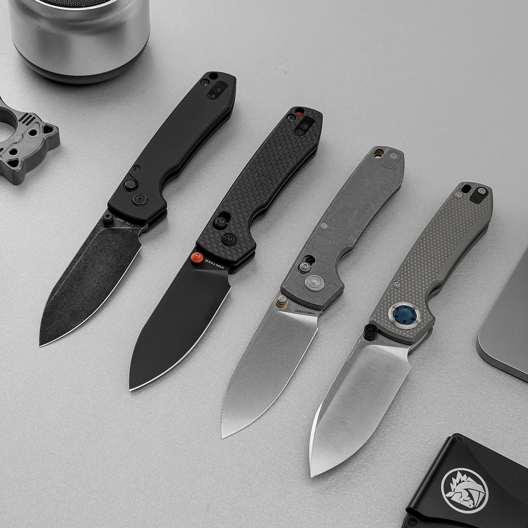 Row of four folding knives with textured handles on a gray surface, blades exposed from left to right, bottom right shows a black case with a logo.
