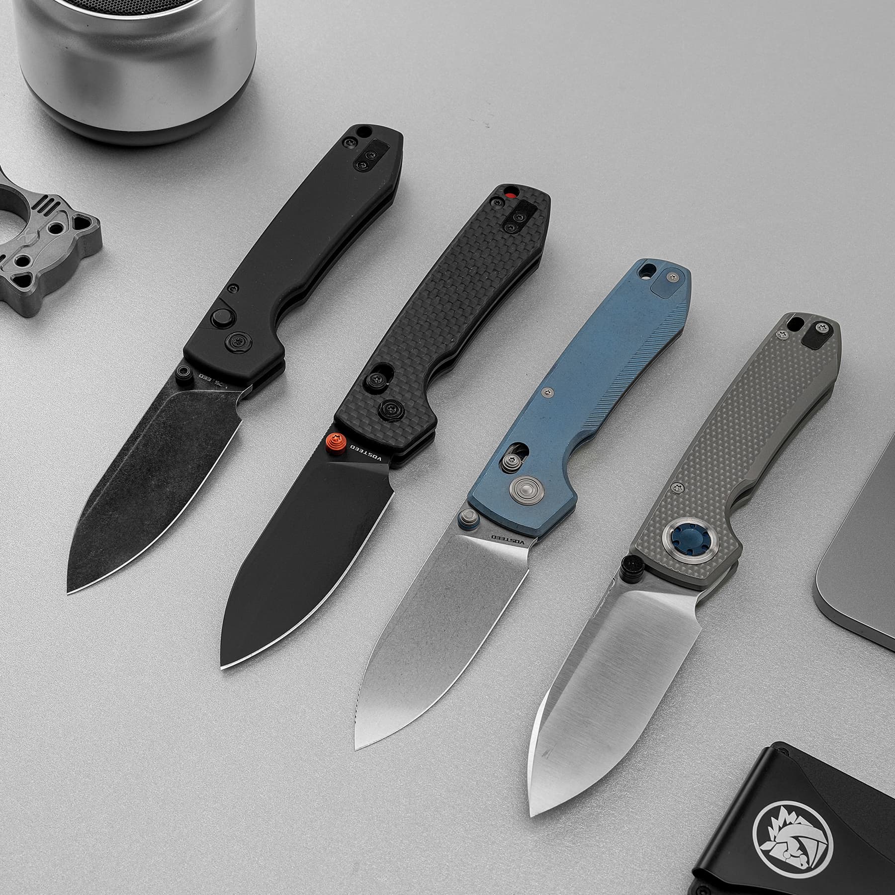 Four folding pocket knives with colored handles (black, blue, gray) and exposed blades laid diagonally on a gray surface.