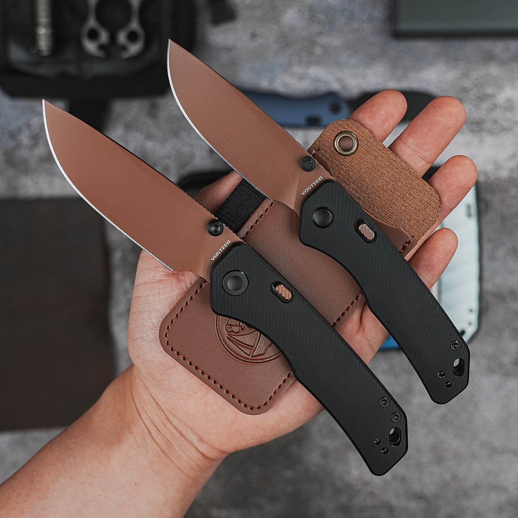 Two folding knives with brown handles and black grips held in a hand over a brown leather sheath.
