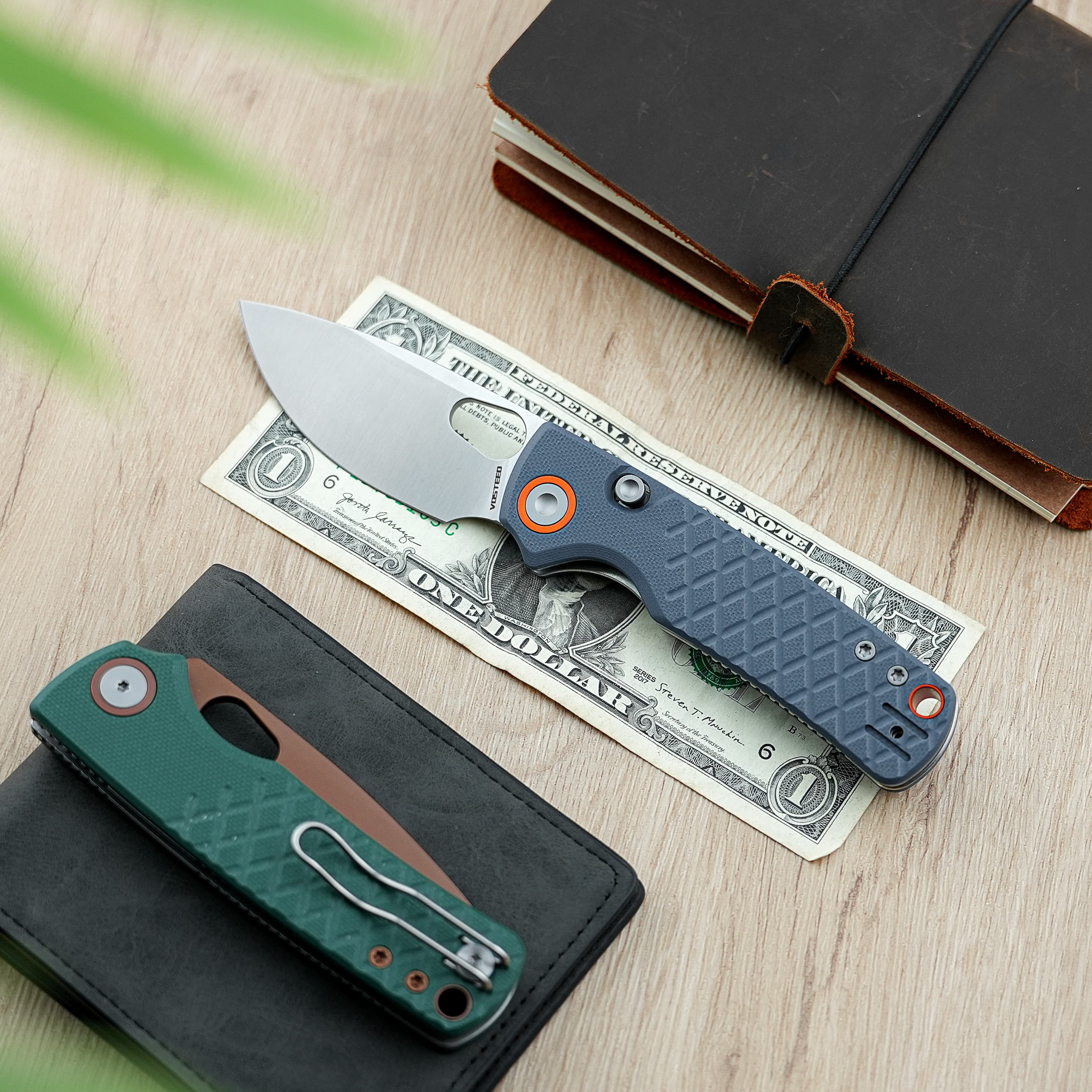 Close-up of a folding knife resting on a one-dollar bill atop a wooden desk near a dark notebook with a leather cover.