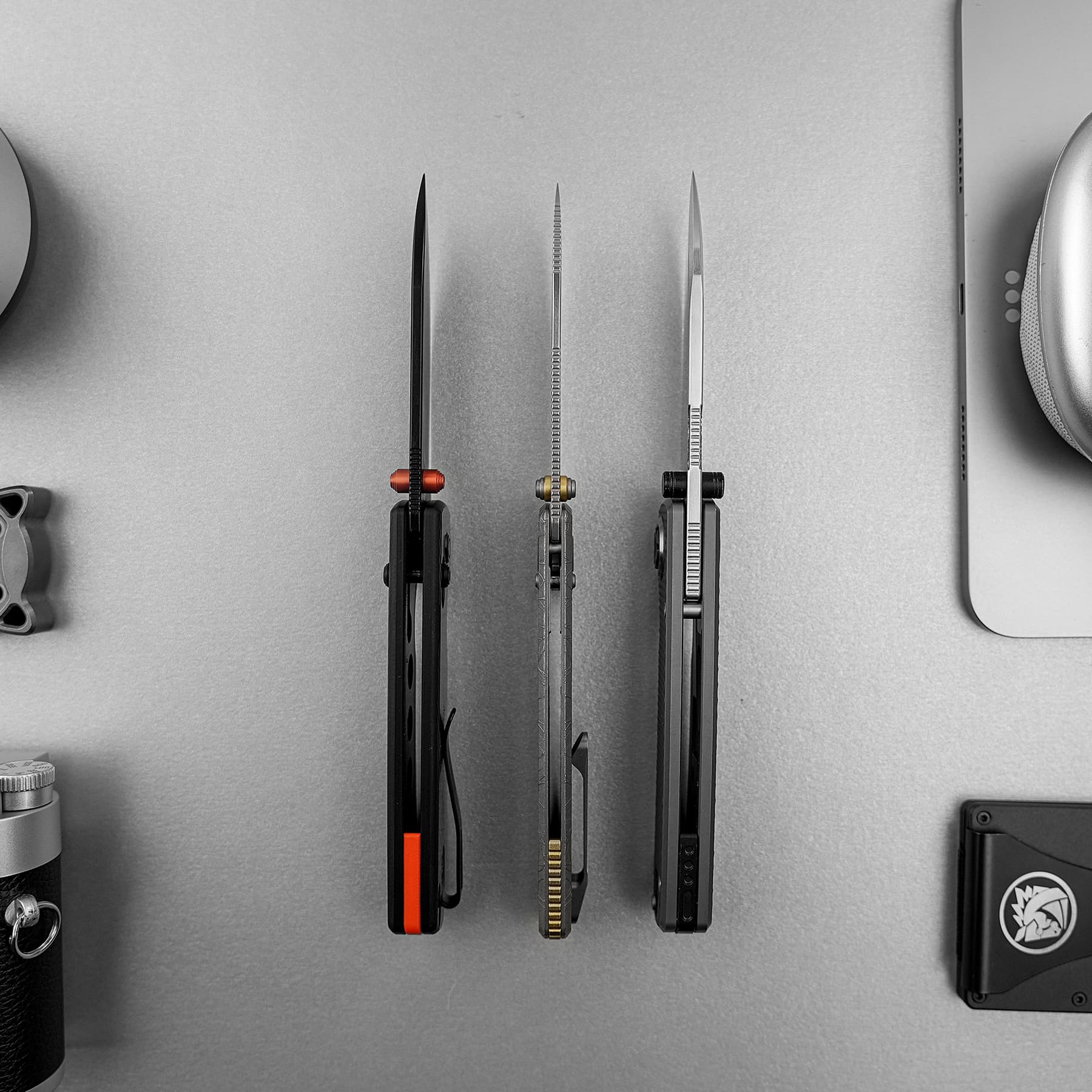 Three precision knives hanging vertically on a gray wall, red, gold, and black-handled tools arranged in a row in a workshop setting.