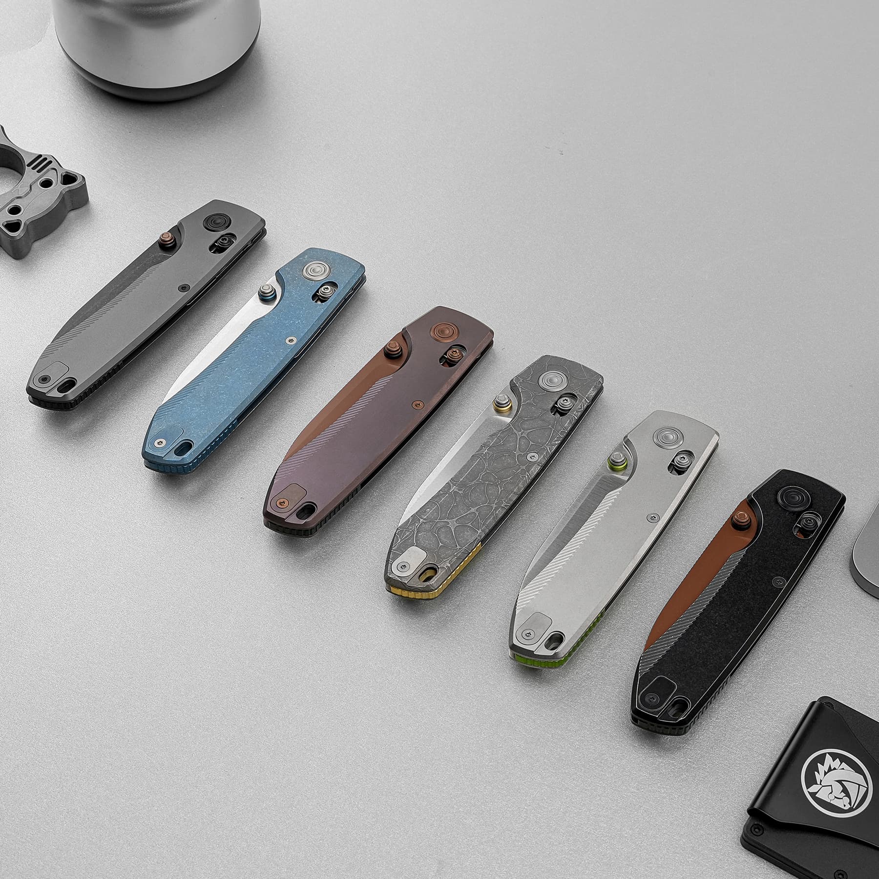 Assorted folding pocket knives laid out diagonally on a gray surface, displaying blue, brown, gray, and marbled handles.