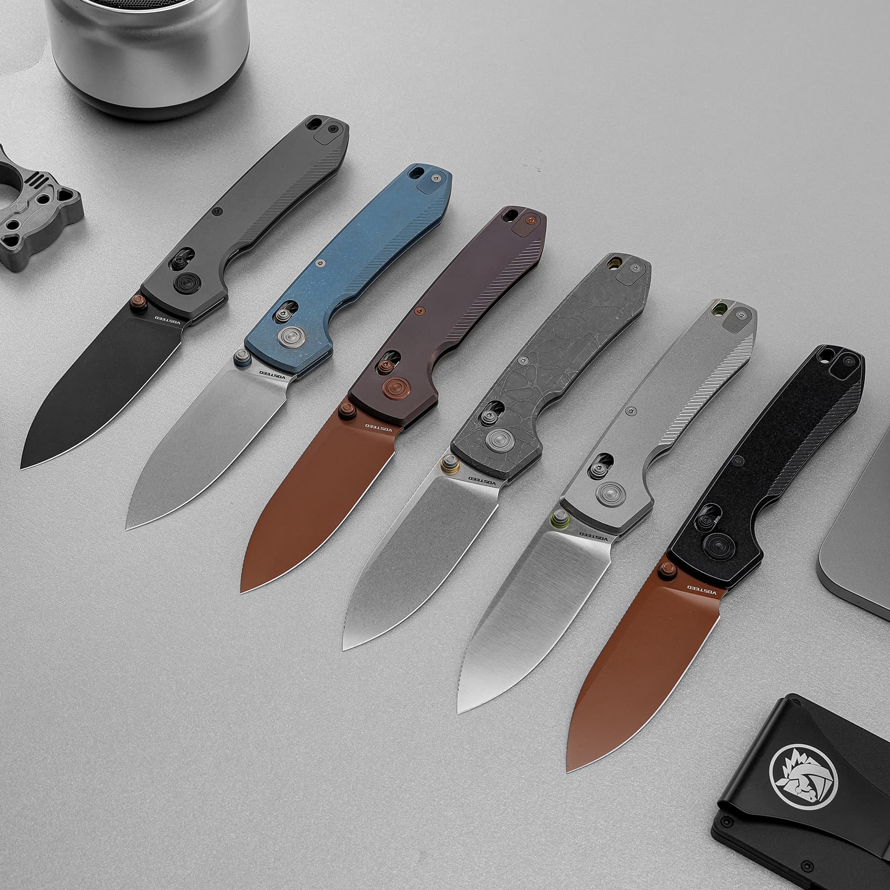 Row of folding knives with angled blades and colored handles laid diagonally on a gray surface.