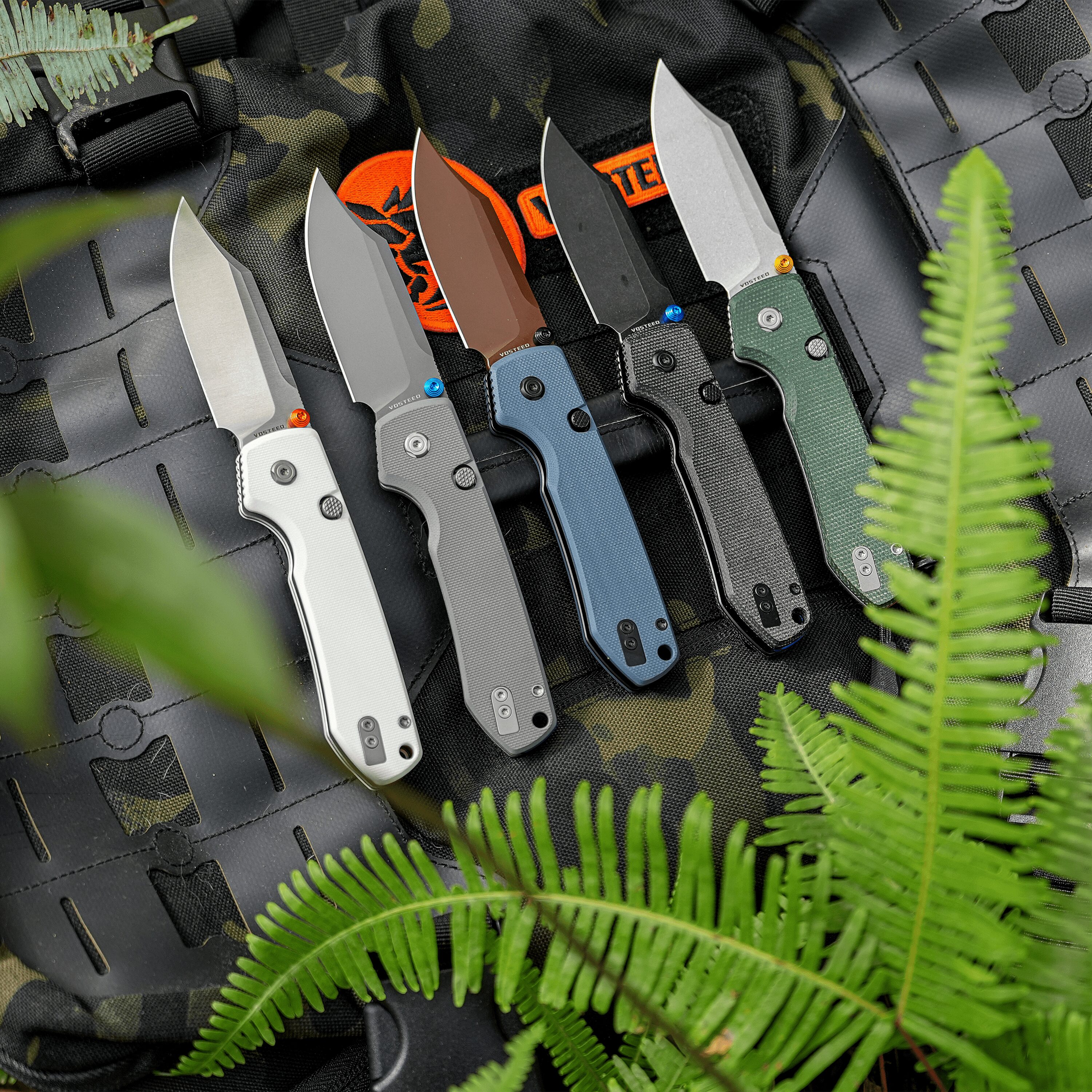 Row of folding knives with colored handles laid in a tactical sheath on camouflage fabric, with green fern fronds in the foreground.