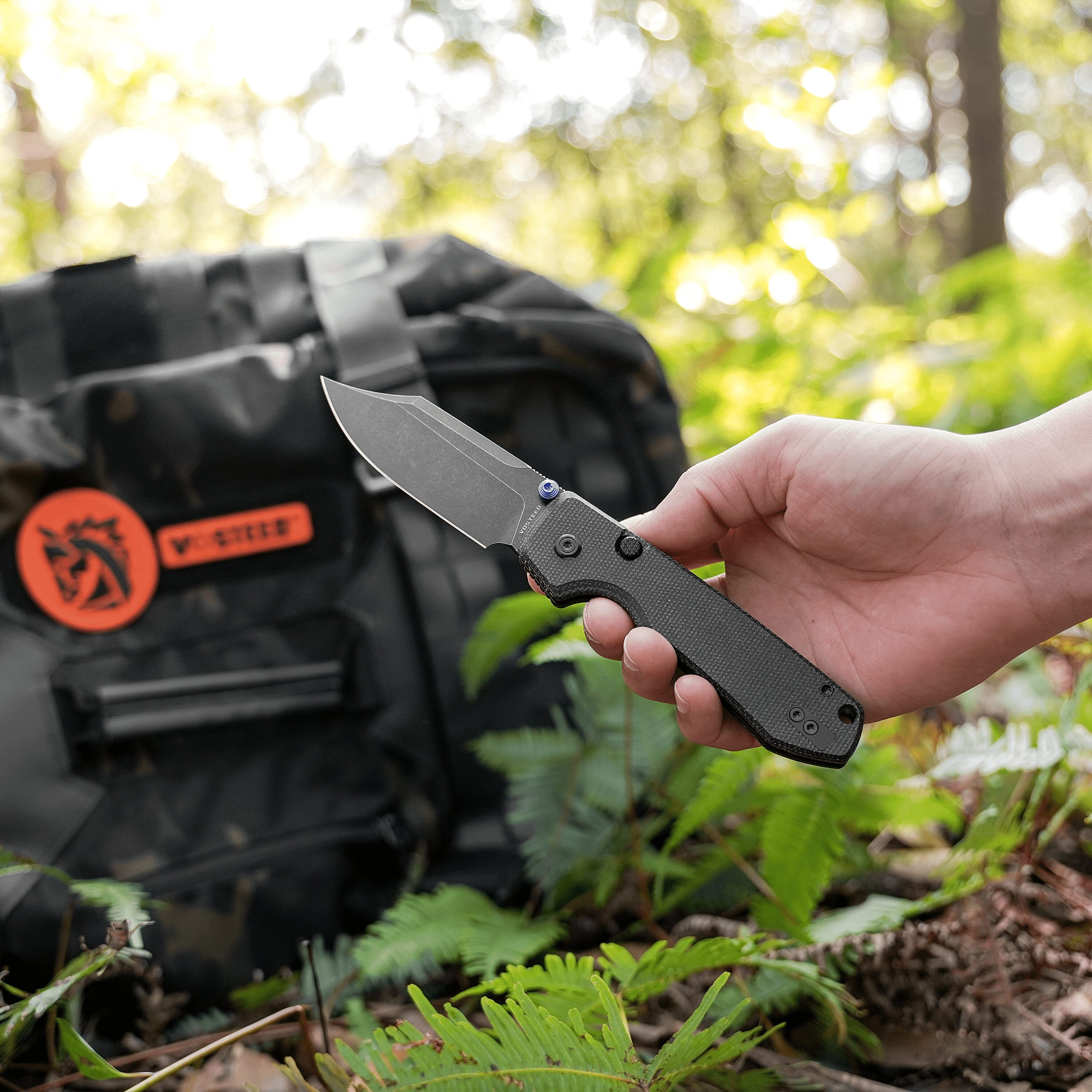 Hand holding a folding knife with a textured black handle, blade exposed, outdoors near a black grill in a wooded setting.
