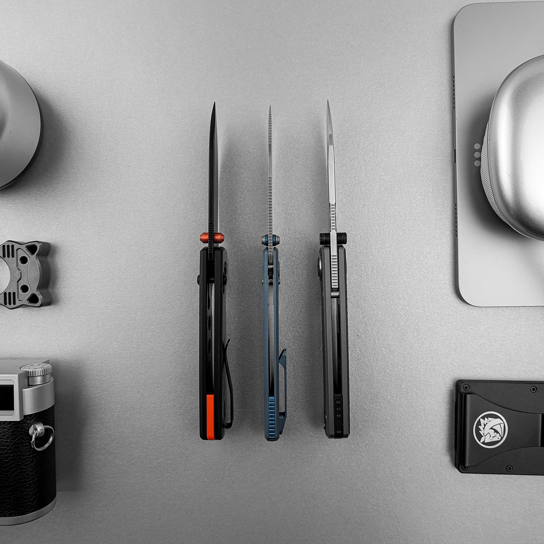 Three precision knives with different blade styles mounted upright on a vertical rack against a gray workbench, with colored grips (orange, blue, black).