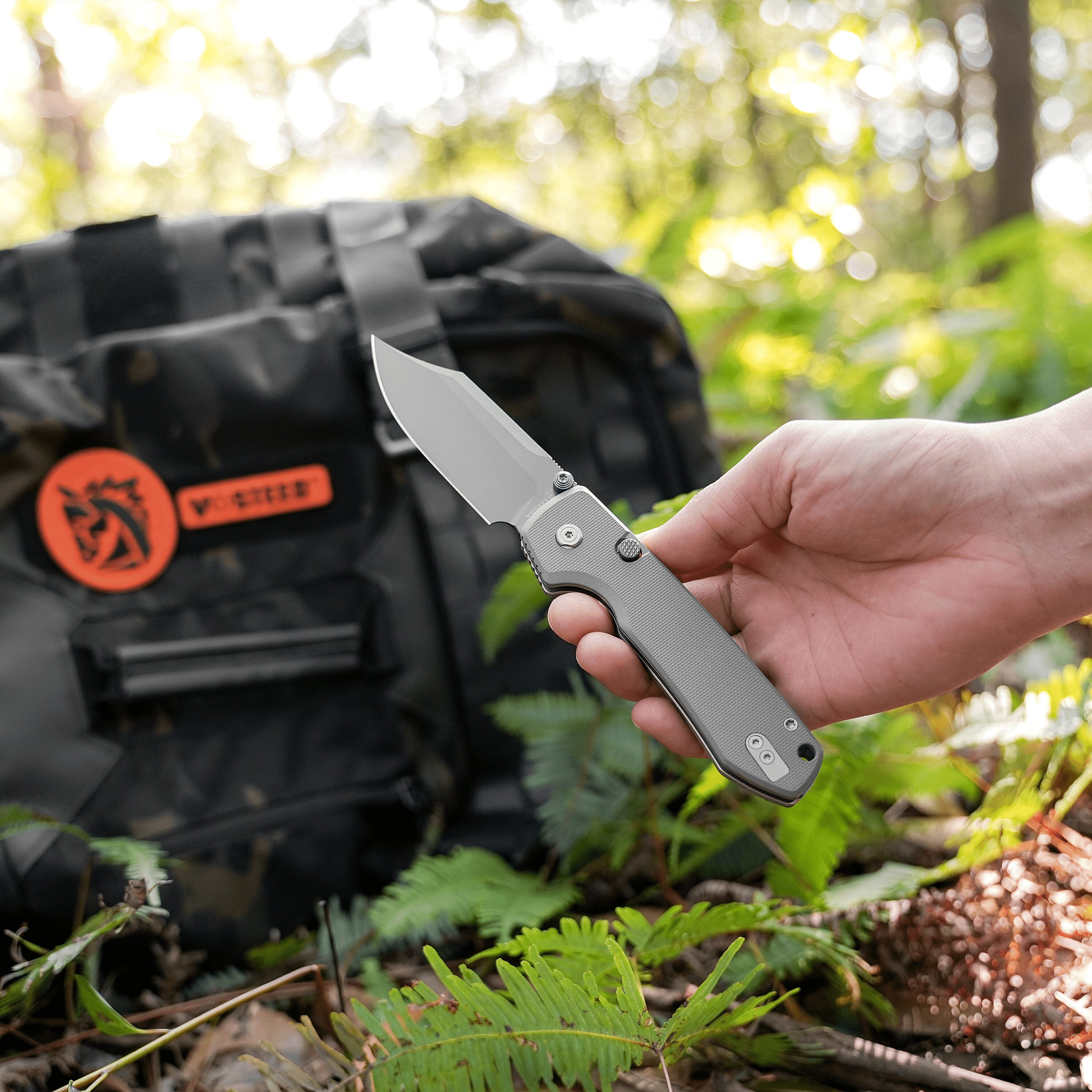 Hand holding a silver folding knife outdoors, with green foliage and a dark backpack blurred in the background.