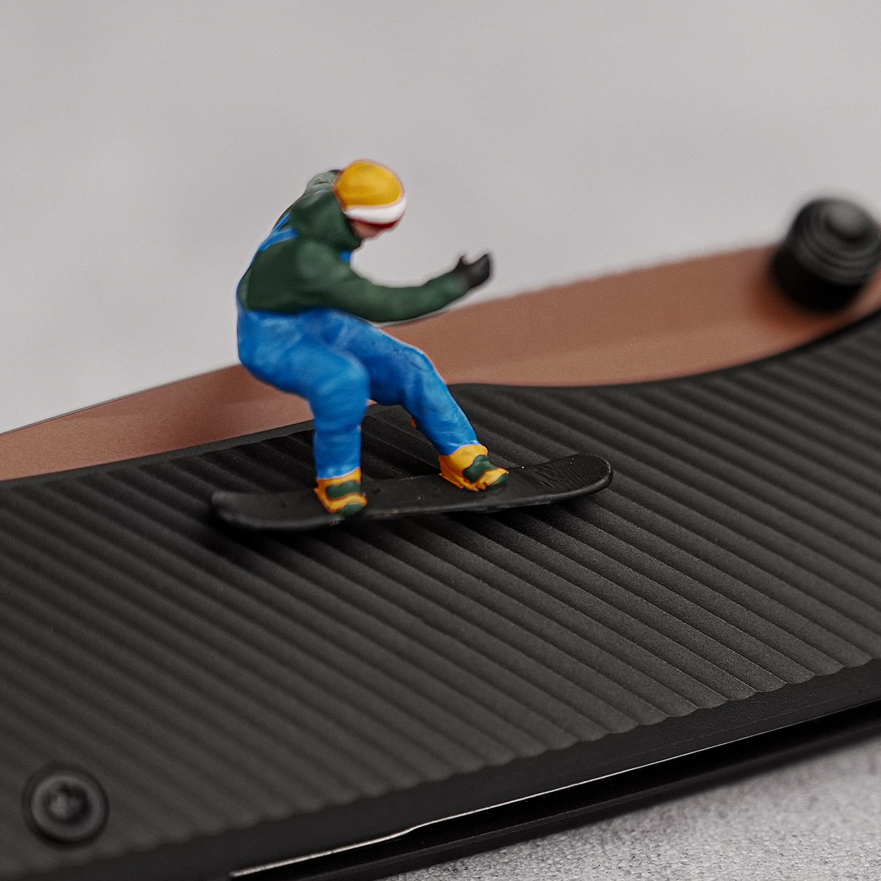 Small plastic action figure in a green jacket and blue pants standing on a tiny black skateboard on a treadmill belt.