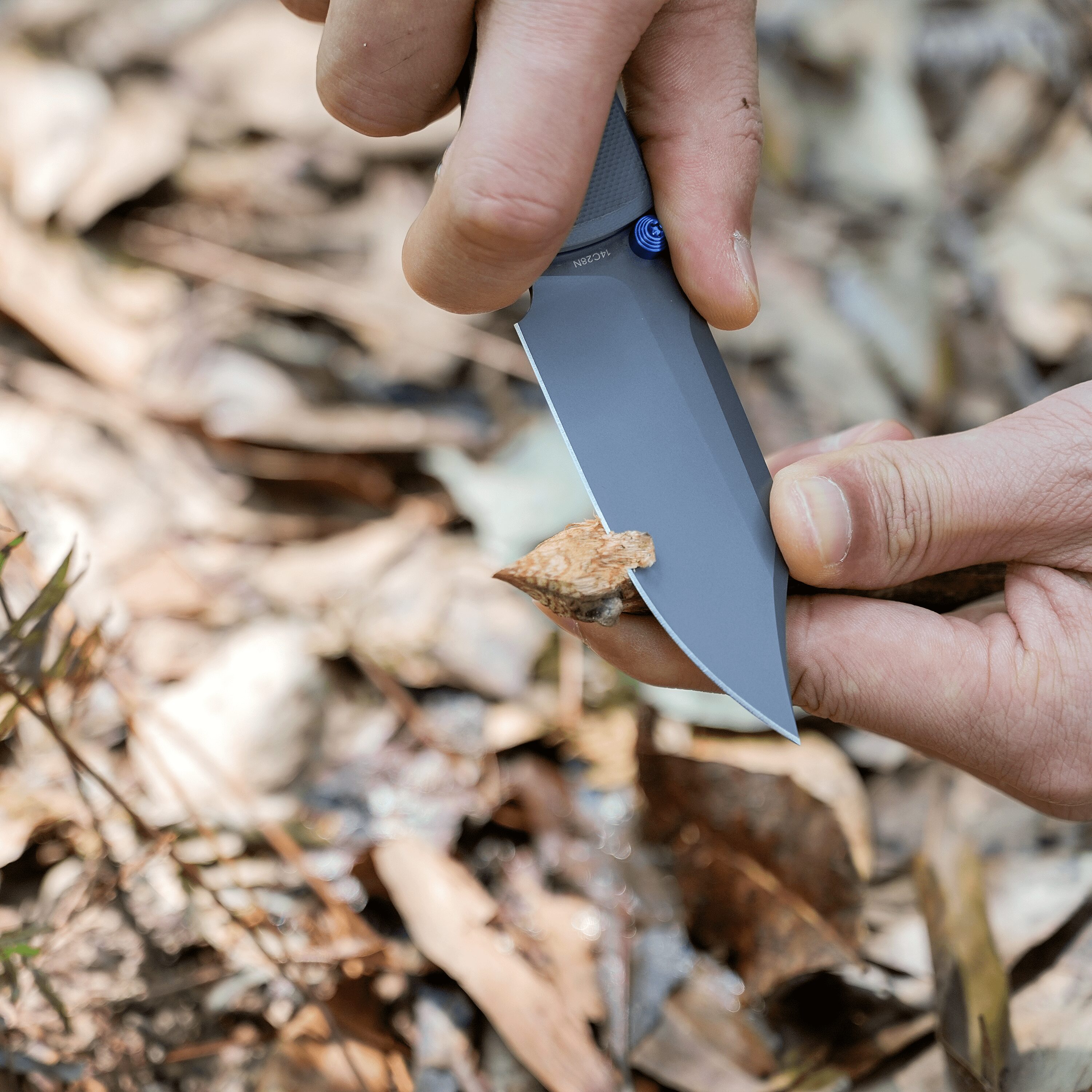 Close-up of hands using a folding utility knife to cut a small piece of wood outdoors.