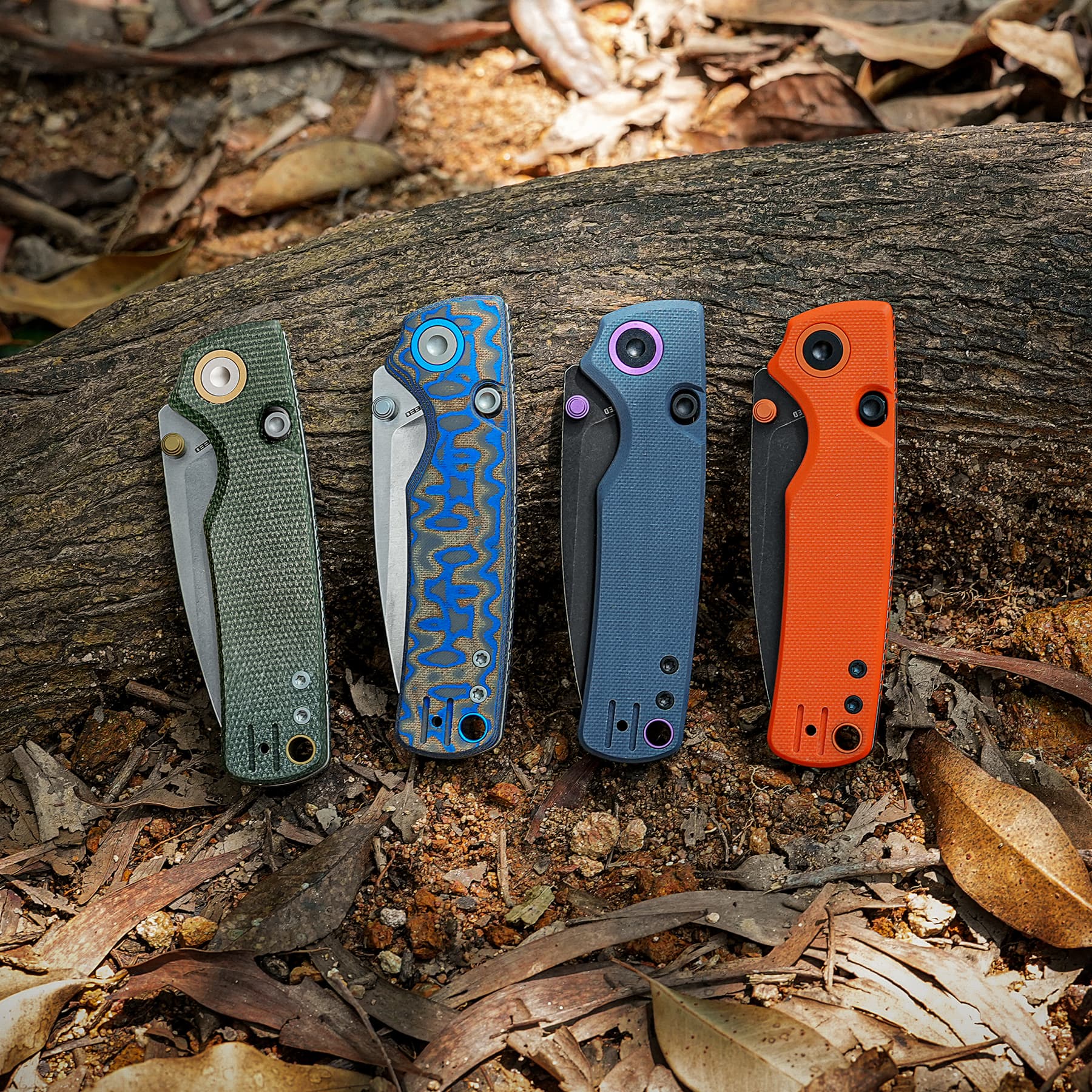 Row of five folding knives with patterned handles laid along a rough log outdoors.