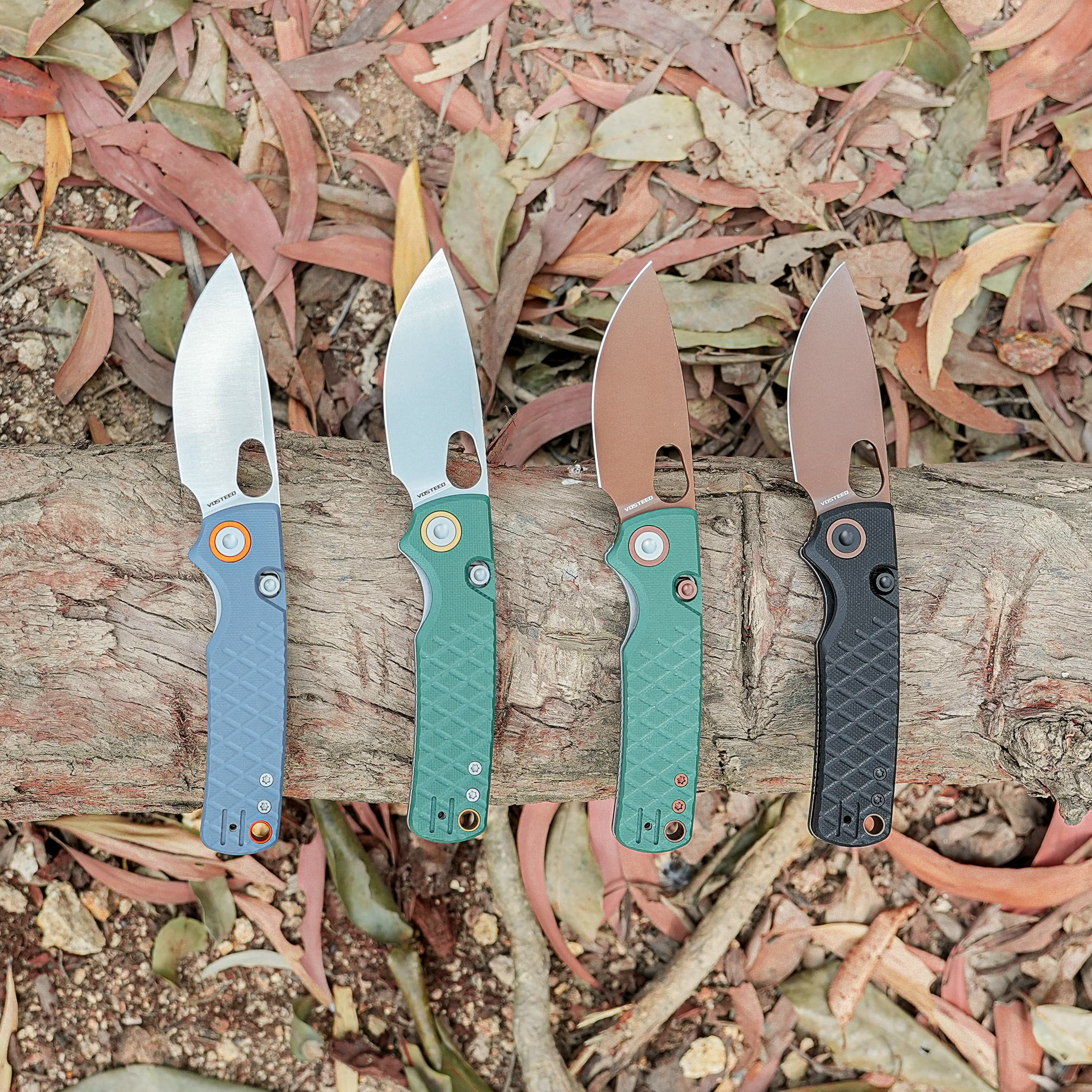 Row of five fixed-blade knives with colored handles laid along a log, against a background of dry leaves and wood.