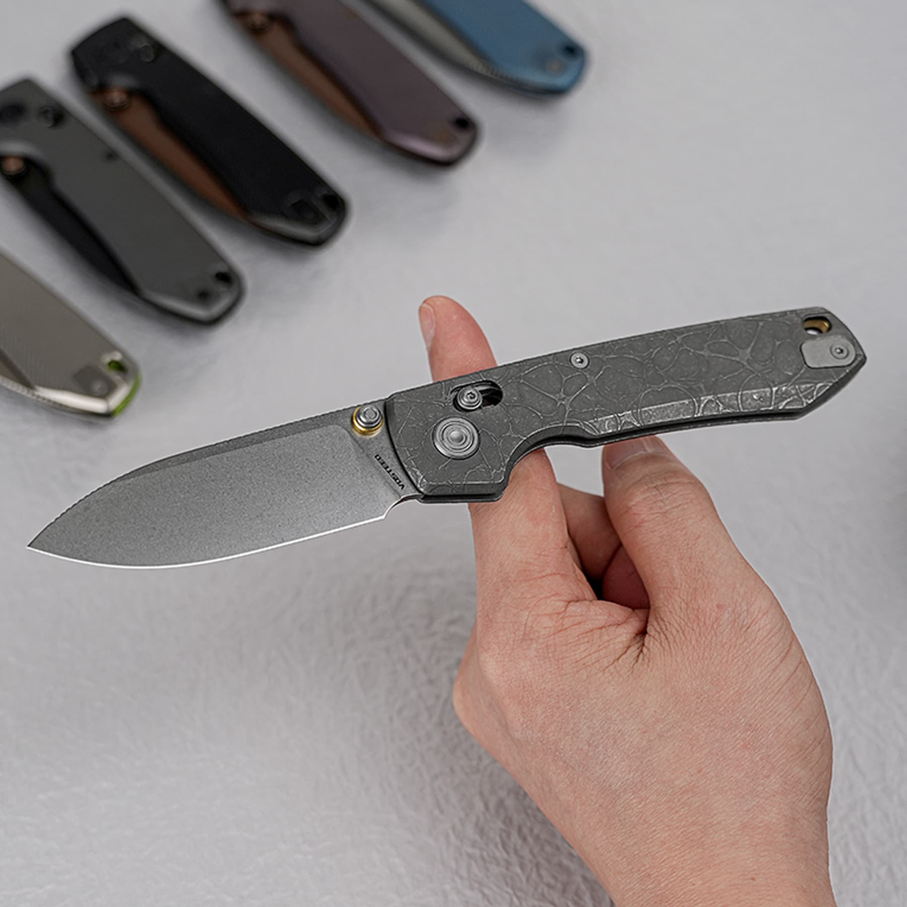 Close-up of a person holding a textured folding knife blade with other colored knives blurred in the background.
