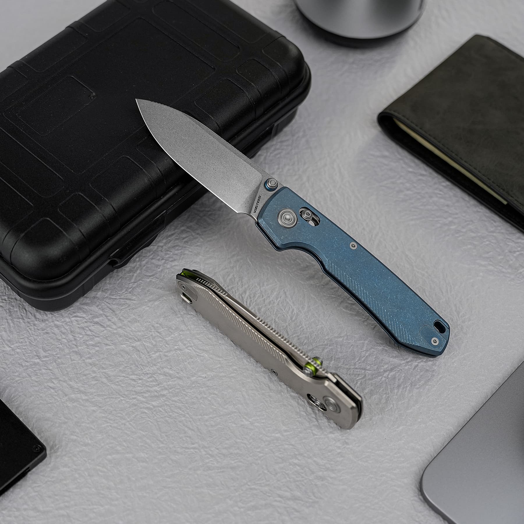 Two folding knives on a textured gray surface with a black hard case and a dark wallet nearby, blade extended from the blue handle.