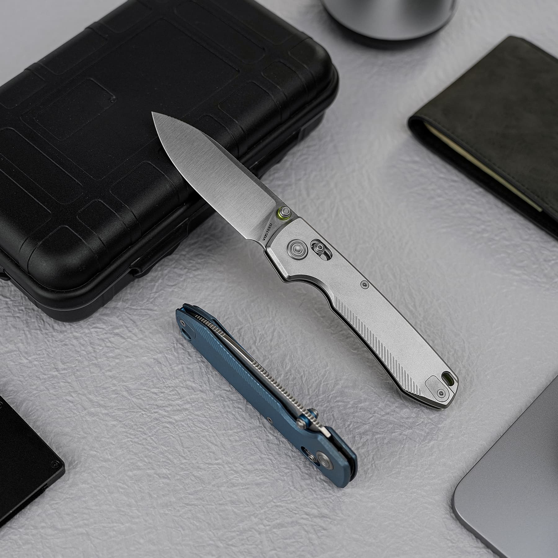Gray folding knife with an open blade resting on a black hard case, with a blue utility knife nearby on a textured surface.