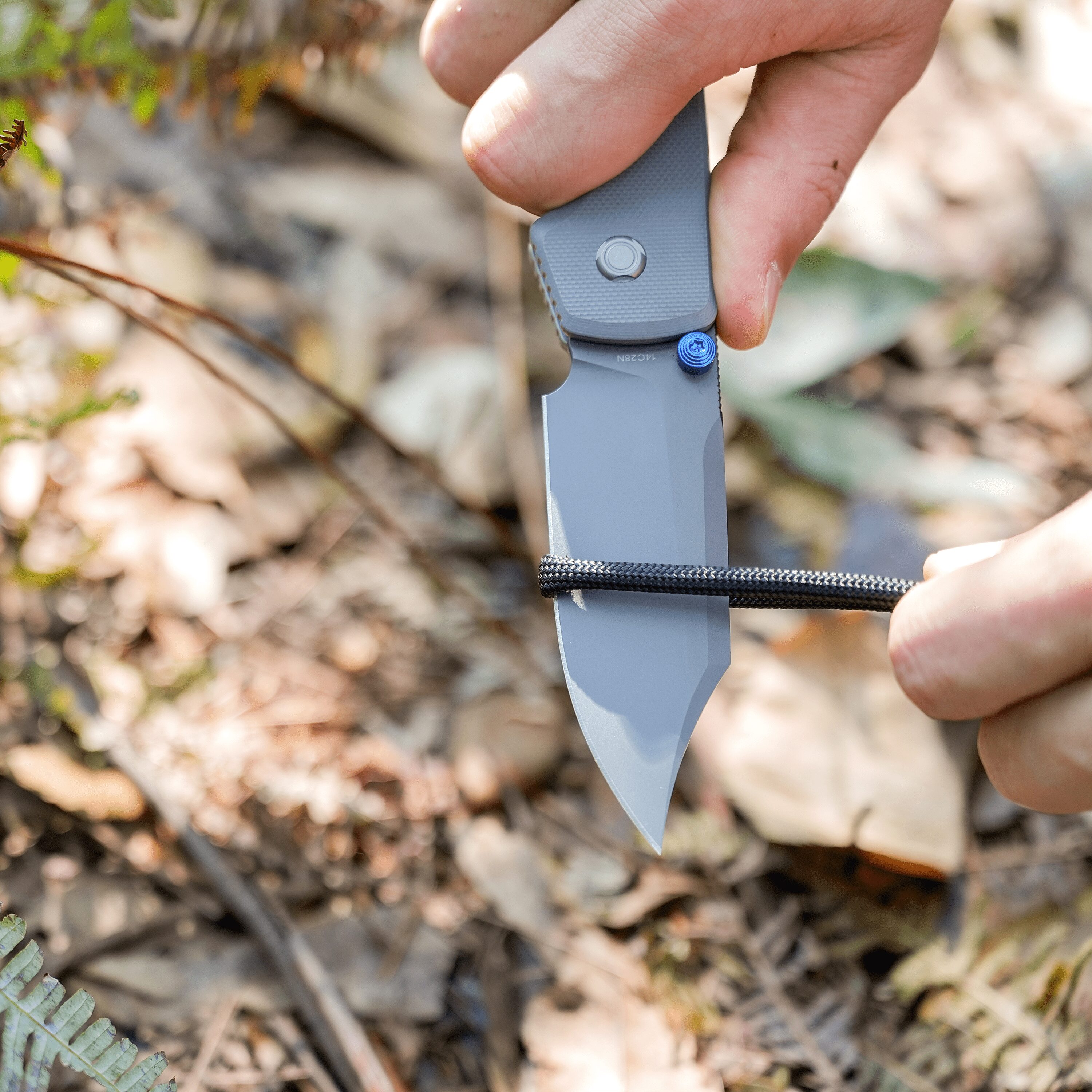 Close-up of a person's hand gripping a folding utility knife outdoors among leaves and twigs.