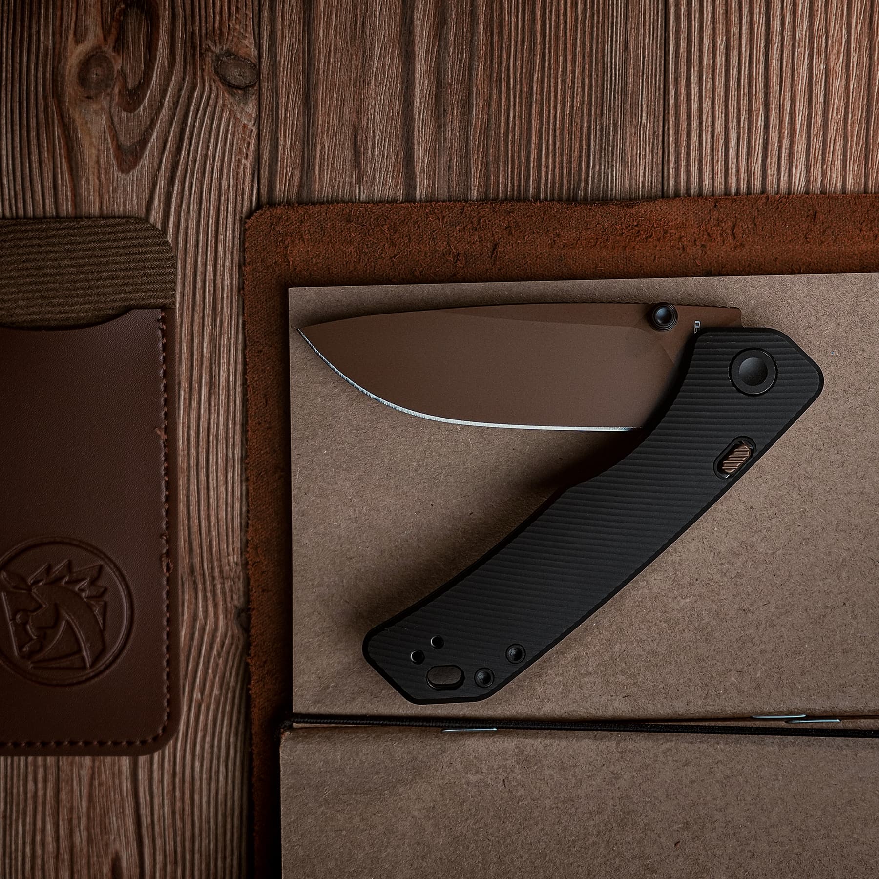 Close-up of a folding knife with a curved blade on a brown leather and wood surface (top-right area).