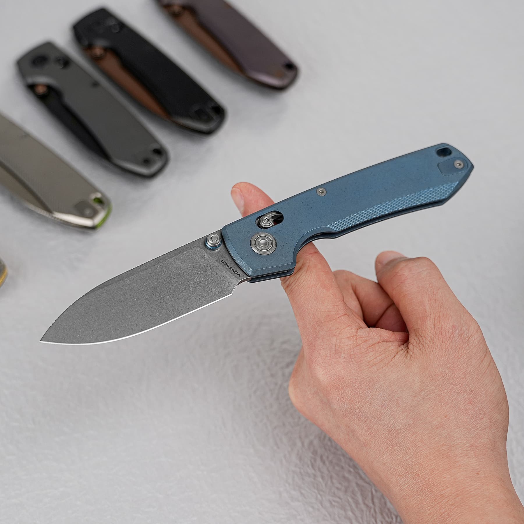 Blue folding knife held in a hand with the blade extended; several other folding knives blurred in the background.