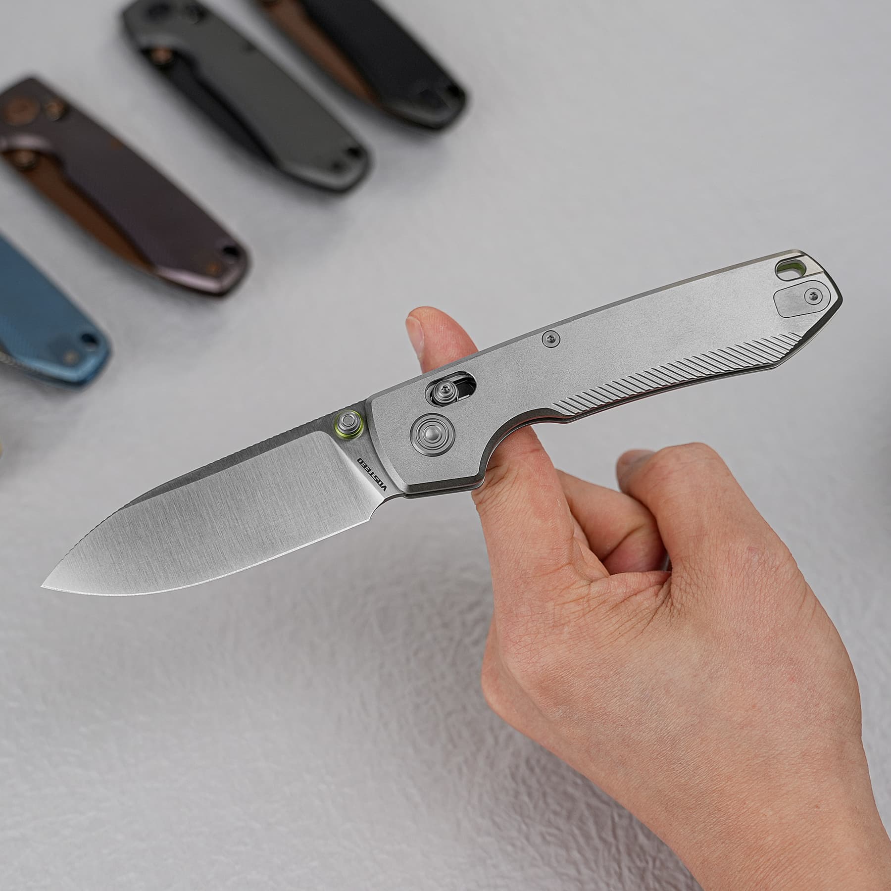 Close-up of a hand holding a silver folding knife, with several other folding knives blurred in the background on a light gray surface.