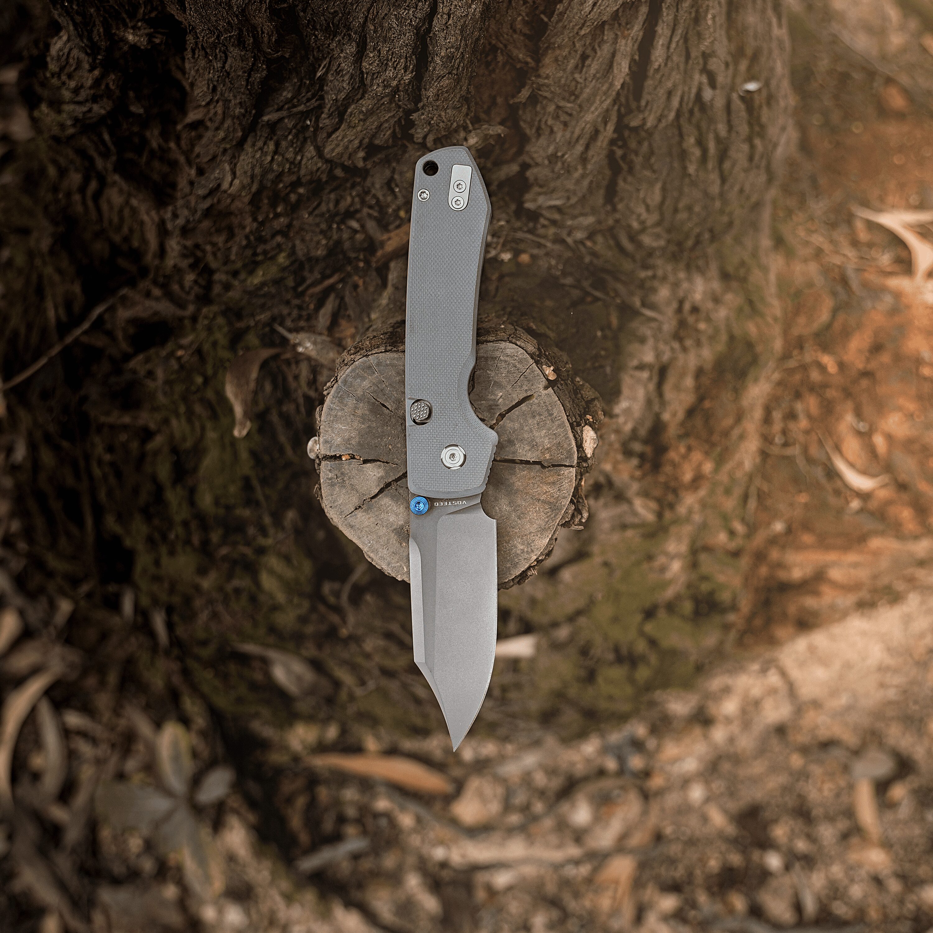 Gray folding knife embedded in a tree stump, blade extended downward and screws visible on the handle.