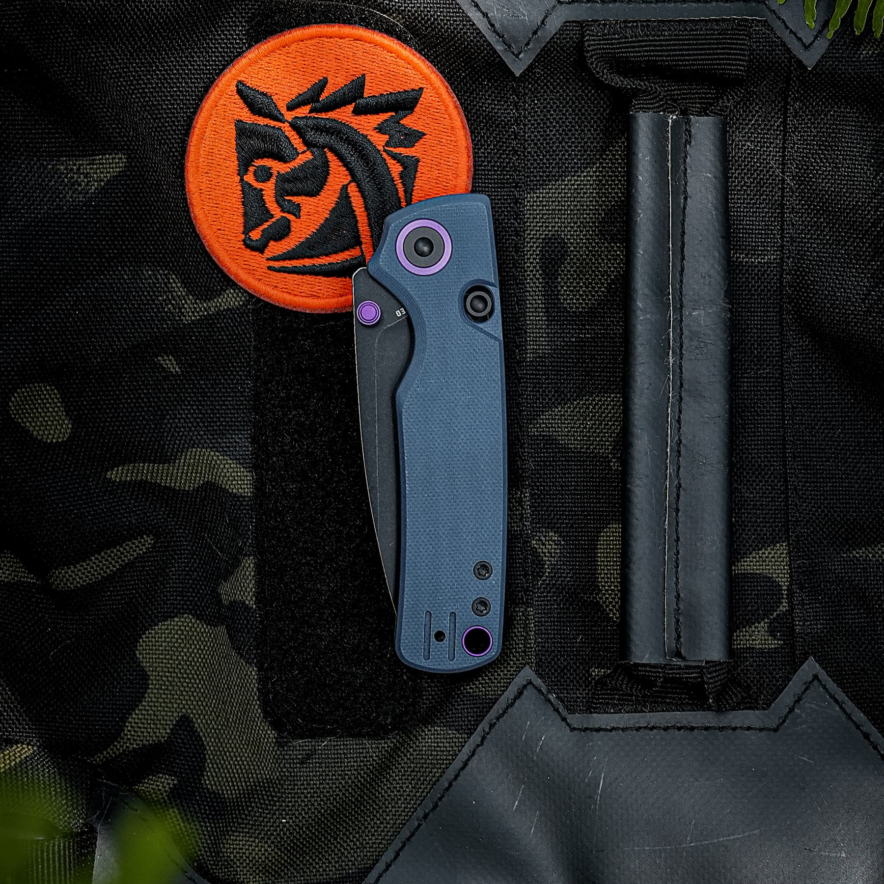 Close-up of a blue folding knife with purple accents resting on a dark camouflage bag beside an orange circular patch with a black emblem.