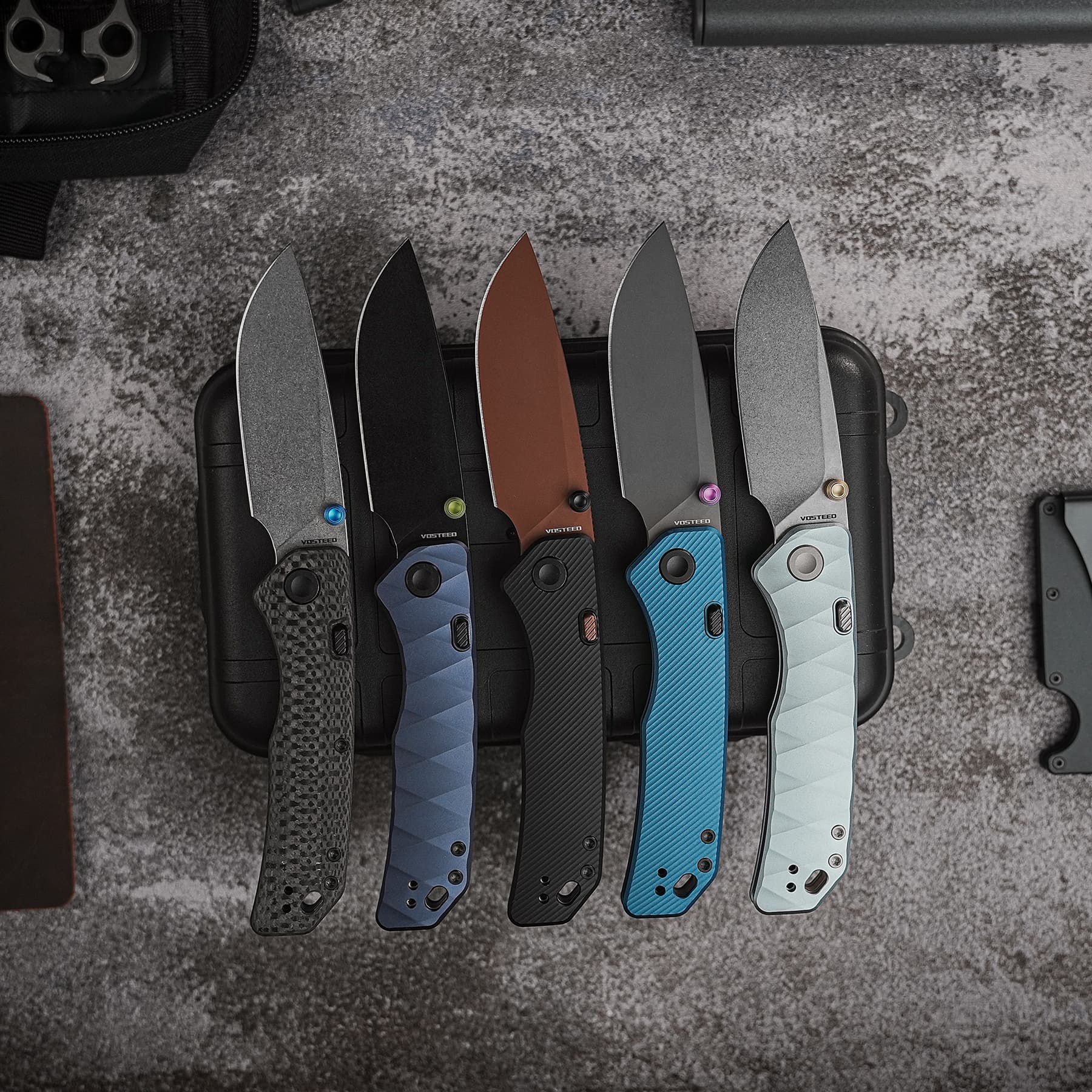 Six folding knives with colored handles arranged in a black case on a textured gray surface, showcasing blue, gray, black, brown, and copper tones.