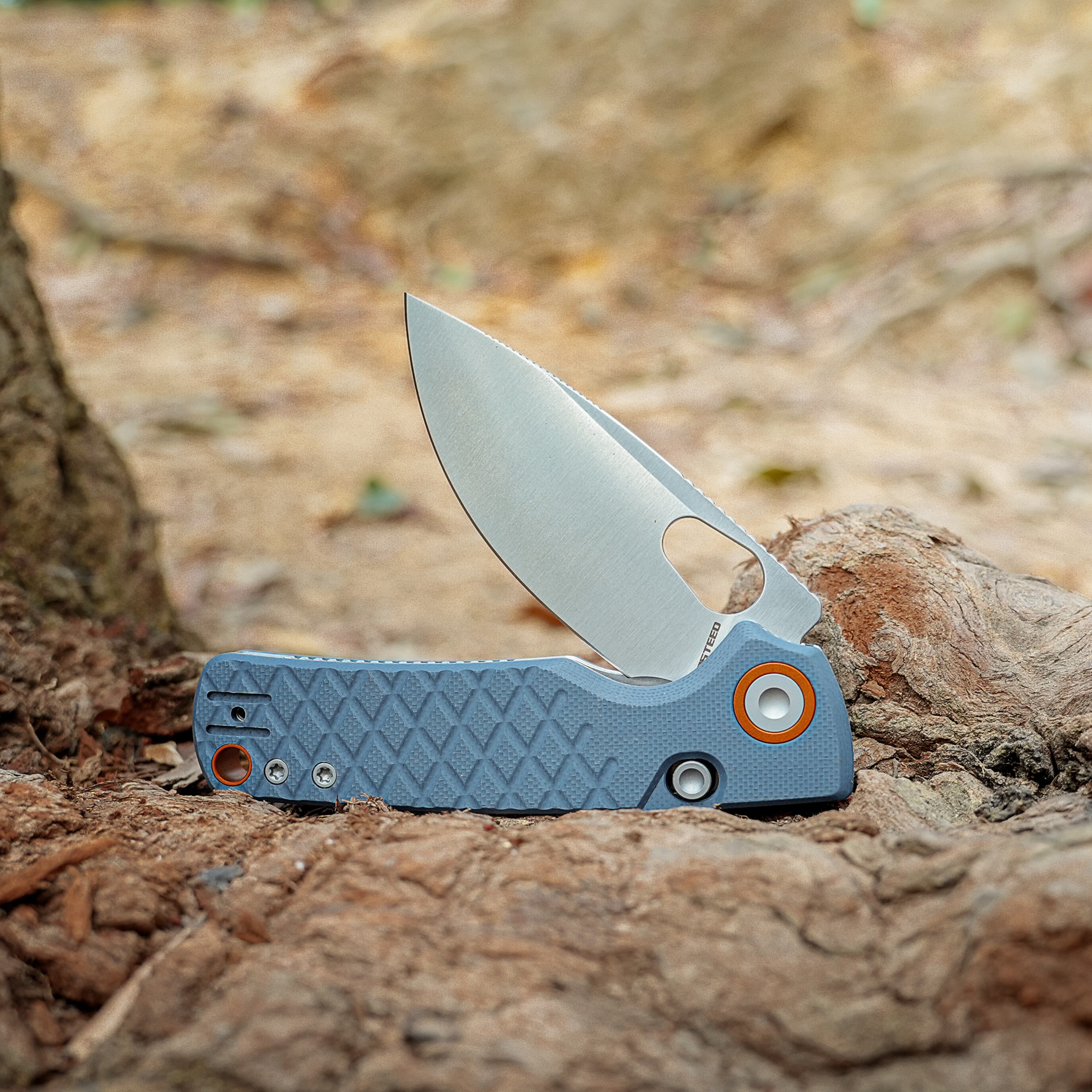 Close-up of a sharp knife blade embedded in a rocky dirt surface, with the blue handle visible in the foreground.