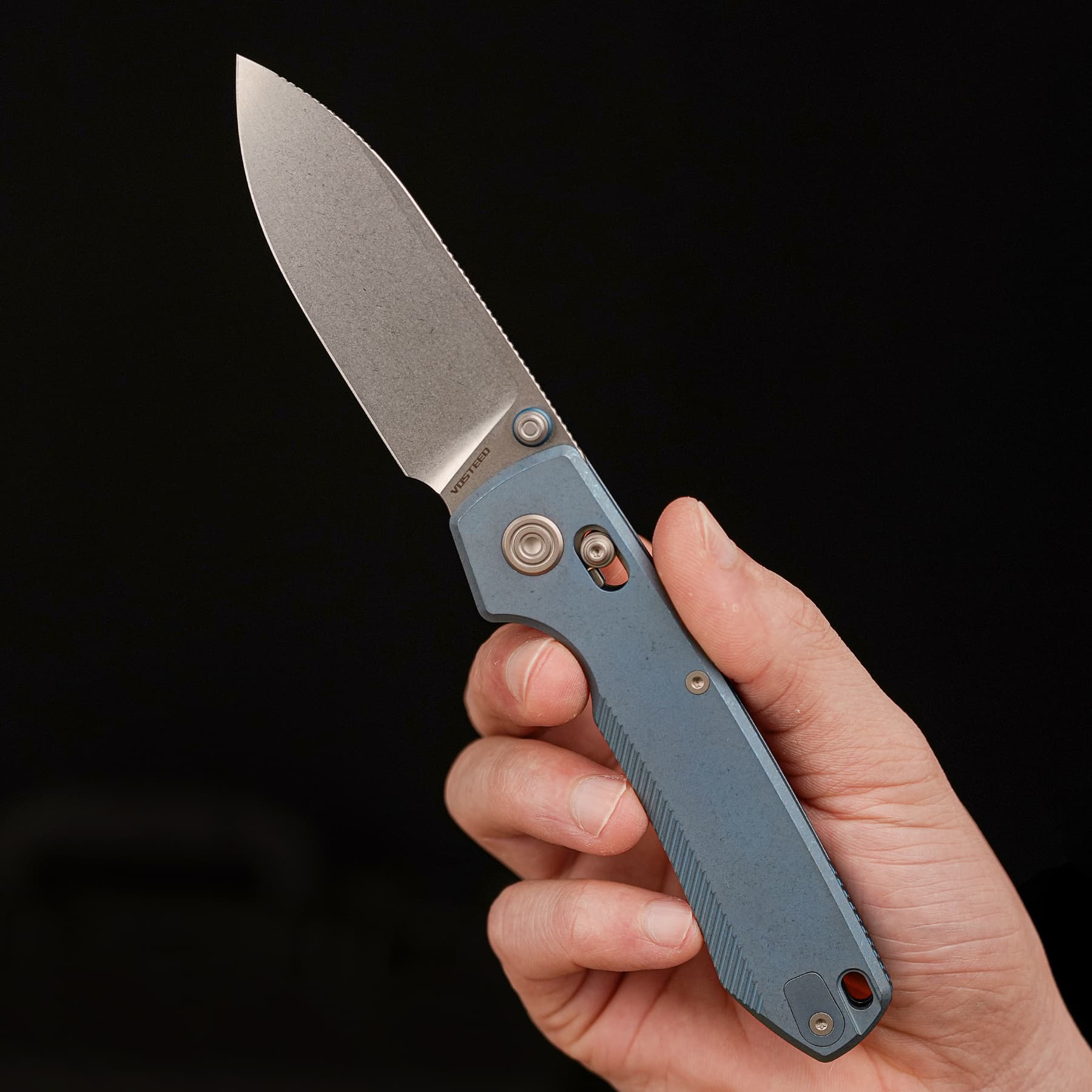 Close-up of a hand holding a grey folding knife with the blade extended against a black background.