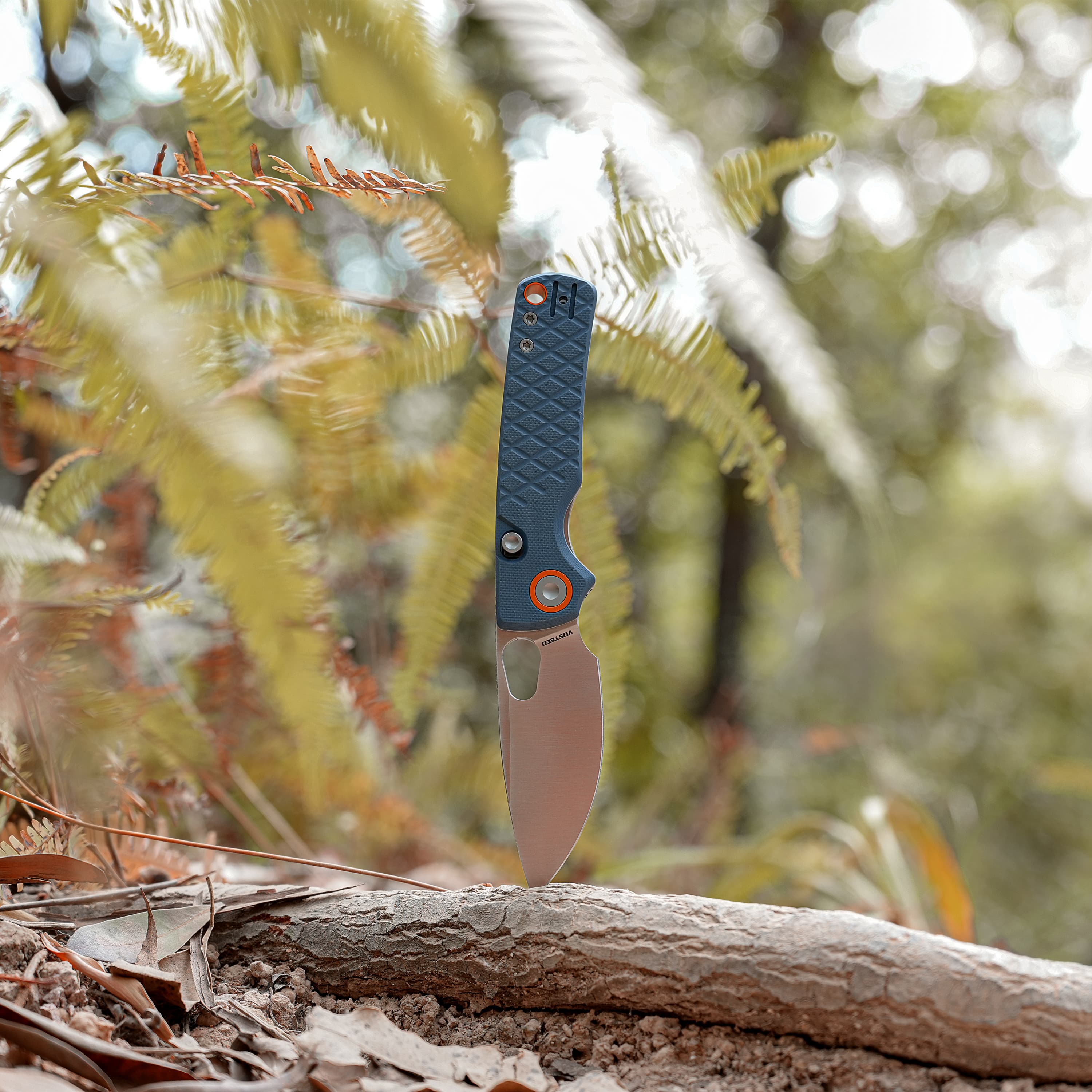 Close-up of a knife blade stabbed into a log outdoors, blue textured handle visible, fallen leaves around.