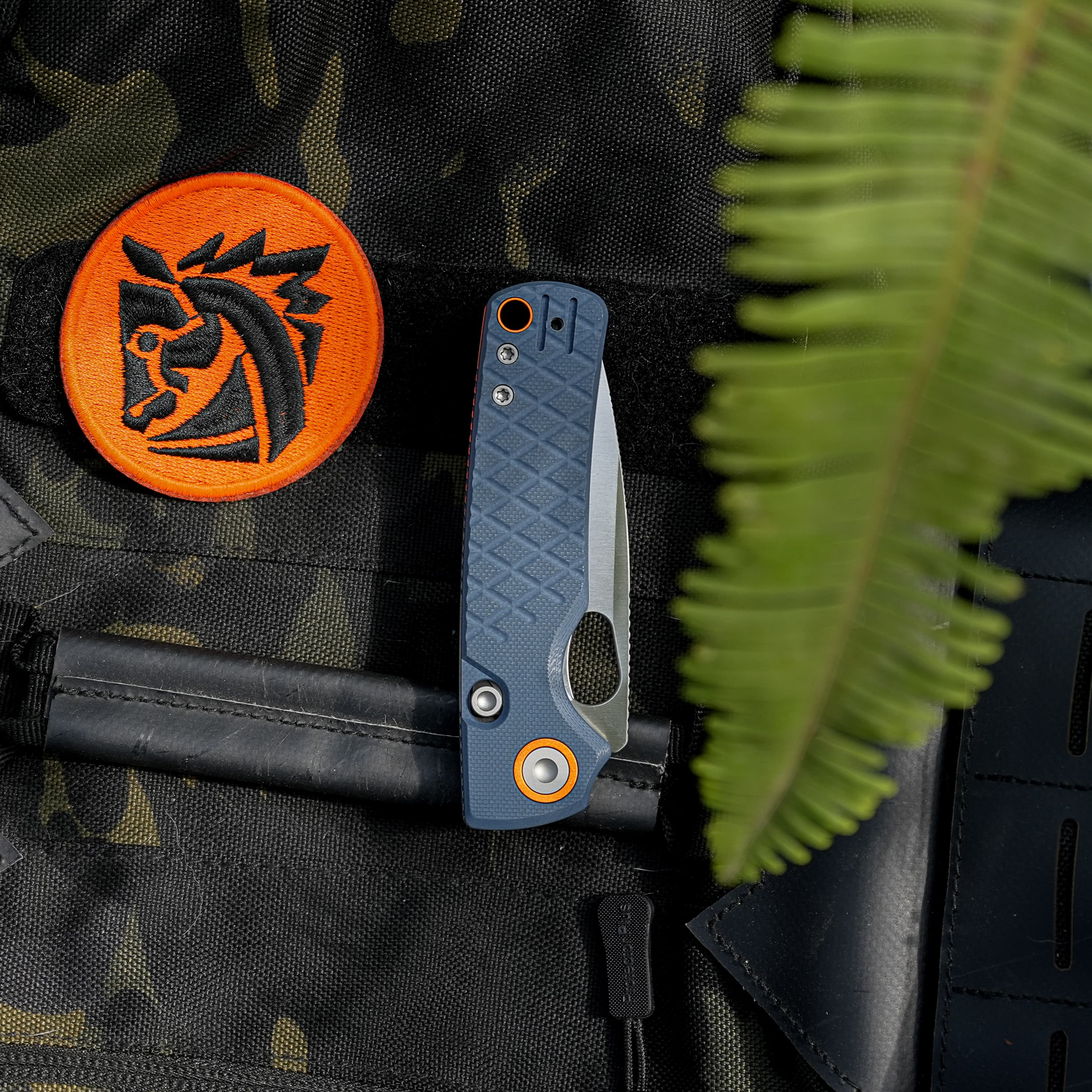 Close-up of camo tactical vest with an orange patch featuring a black emblem, a blue folding knife handle, and a blurred green fern in the foreground.
